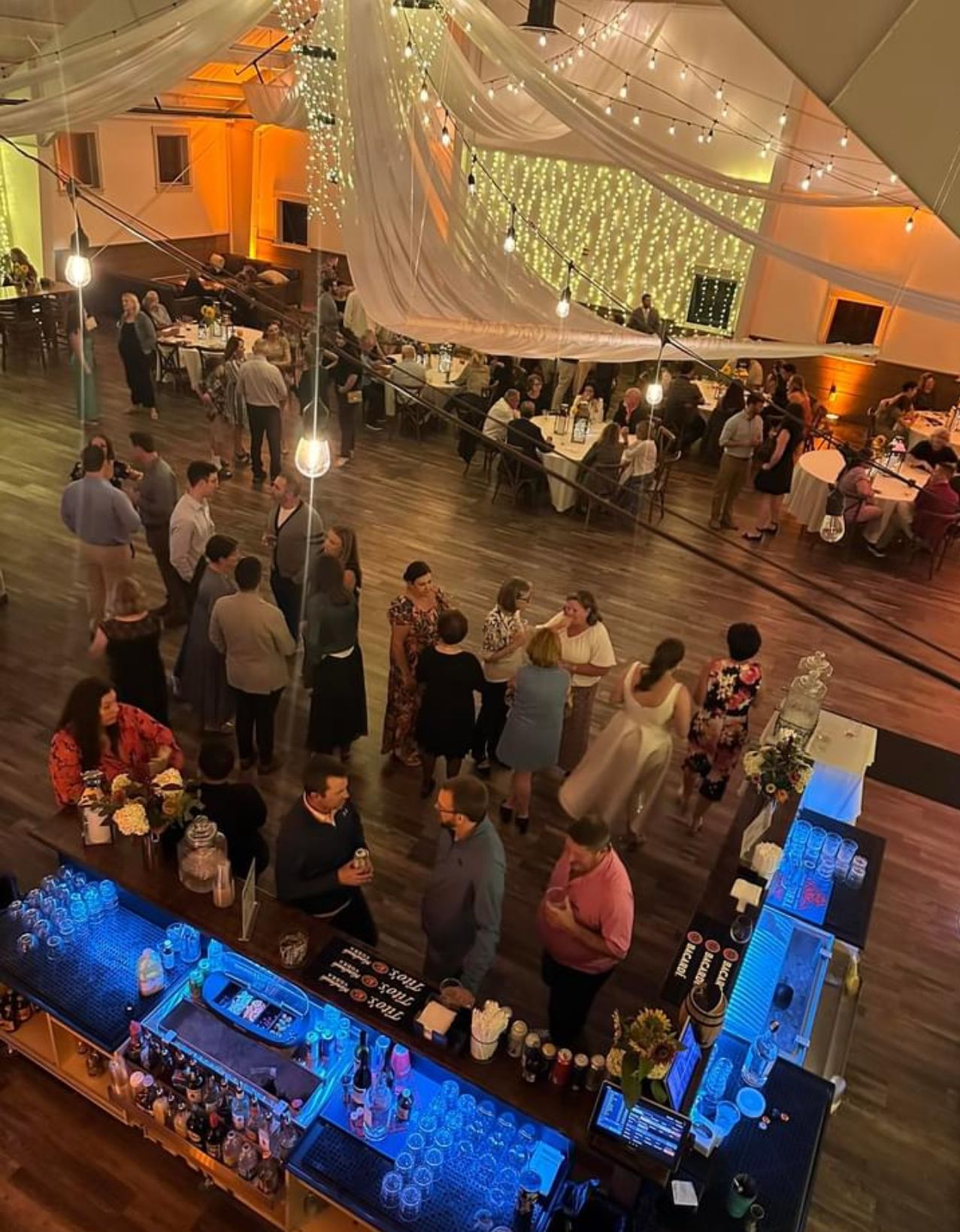 Wedding reception: Guests mingle near a bar, tables set up in a large hall with draped fabric, and fairy lights.