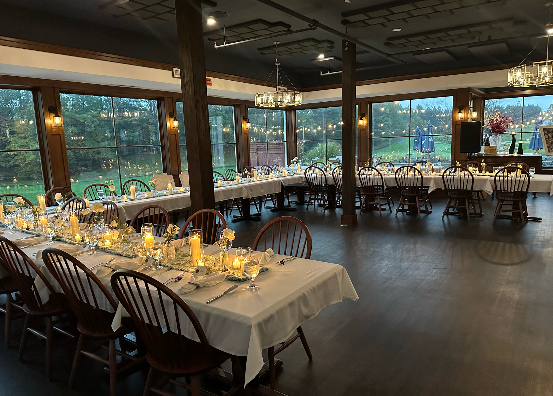 Dining room set for a formal event, with long tables, white linens, candles, and outdoor view.