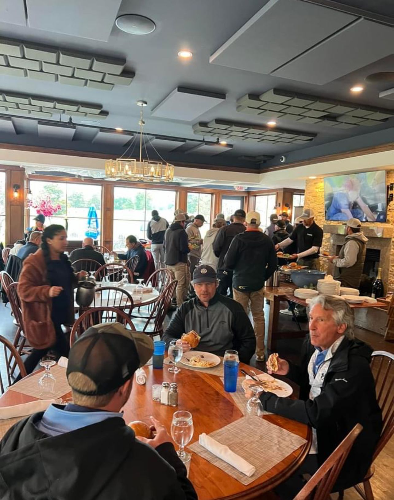 People eating and socializing in a restaurant with large windows. Several tables and chairs. Fireplace, TV, and chandelier present.