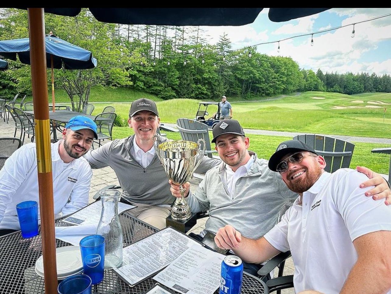 Four men smile, celebrating a golf victory outdoors, one holding a trophy.