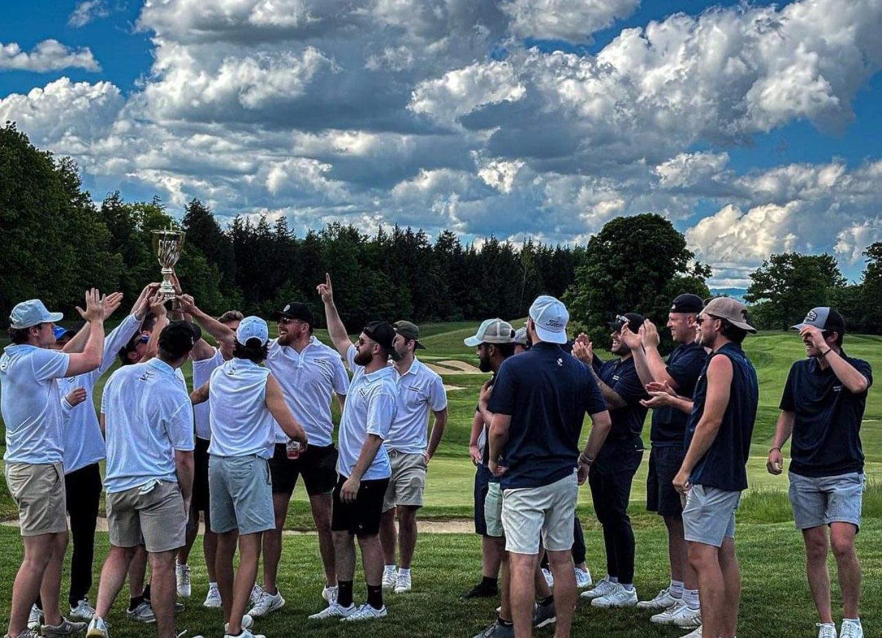 Golf team celebrates win, holding trophy high on green with blue sky.