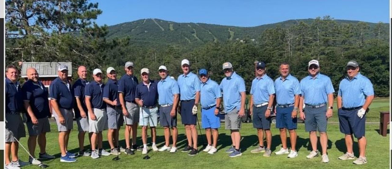 Group of men in golf attire, posing on a green with mountains in the background.