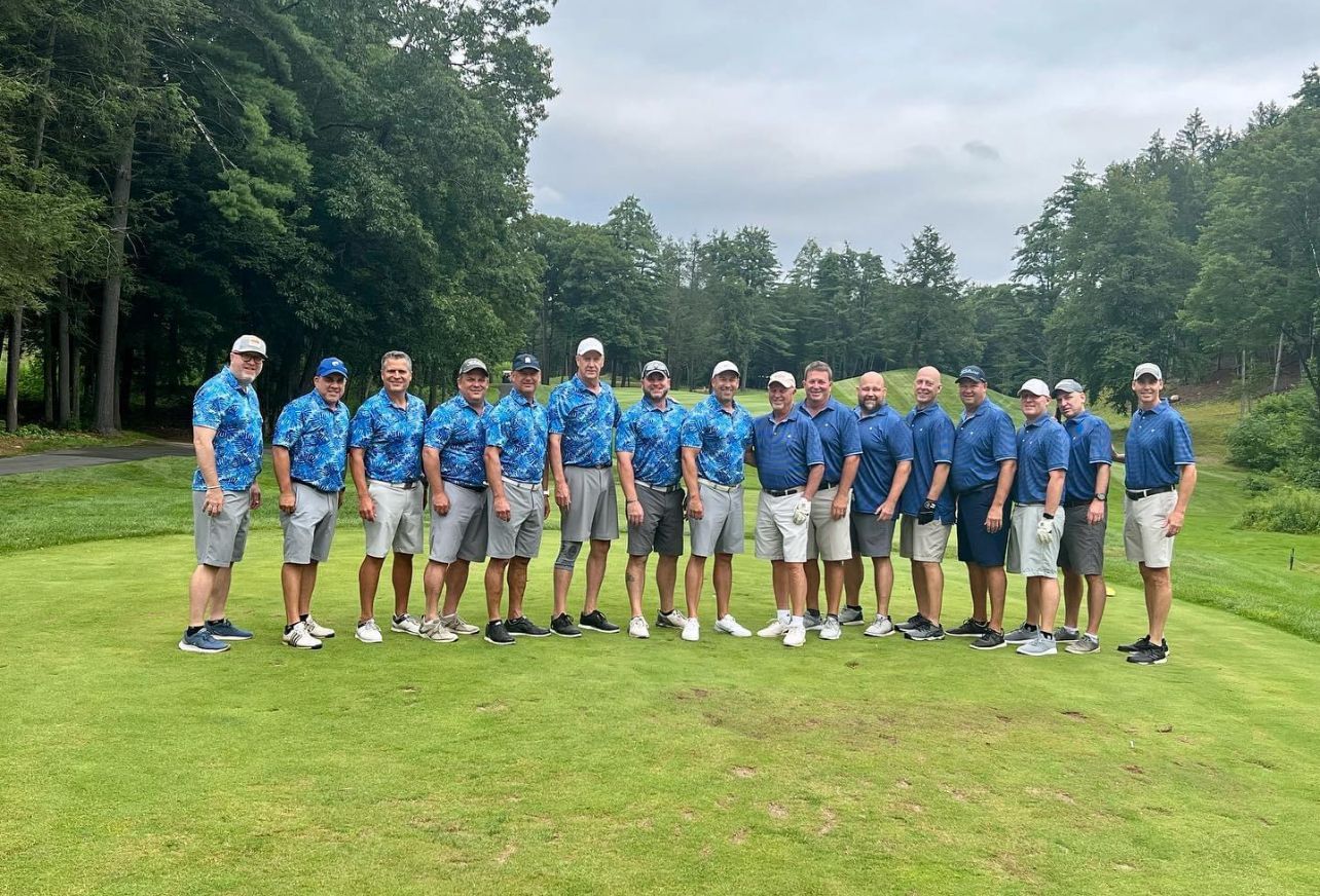 Group of men in blue shirts and gray shorts on a golf course, posing for a photo.