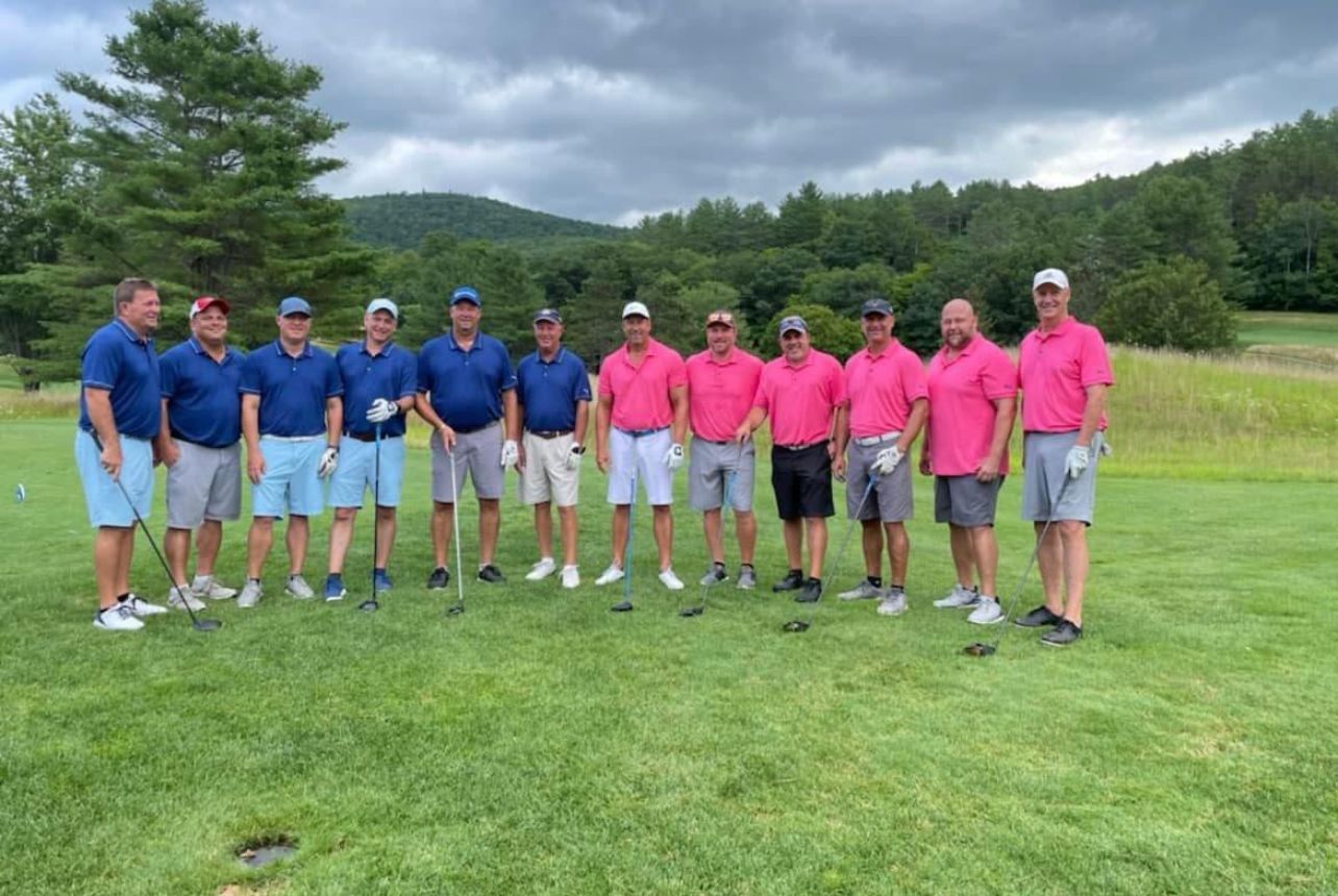 Group of golfers, some in blue shirts, some in pink, posing on a green golf course with mountains in the background.