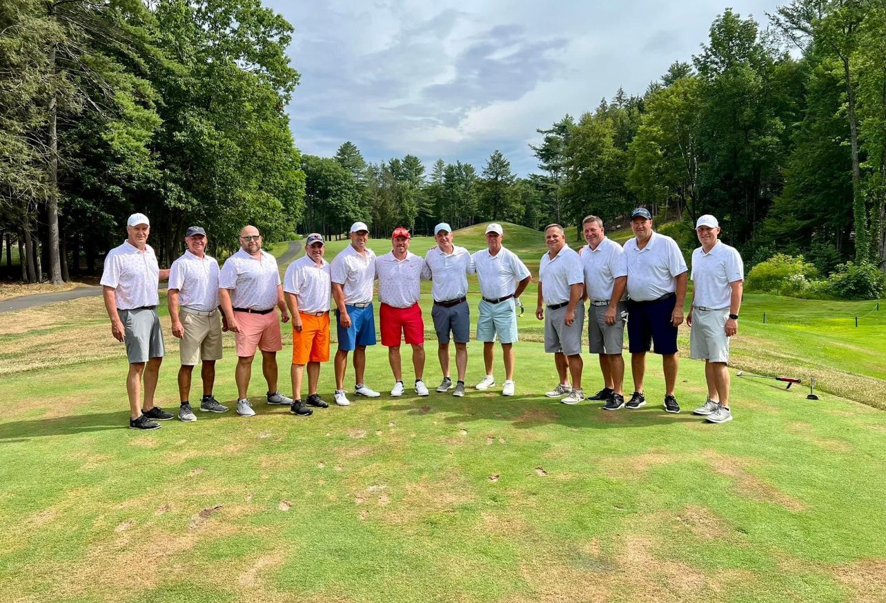 Group of men in golf attire pose on a green, sunny day, with trees in the background.
