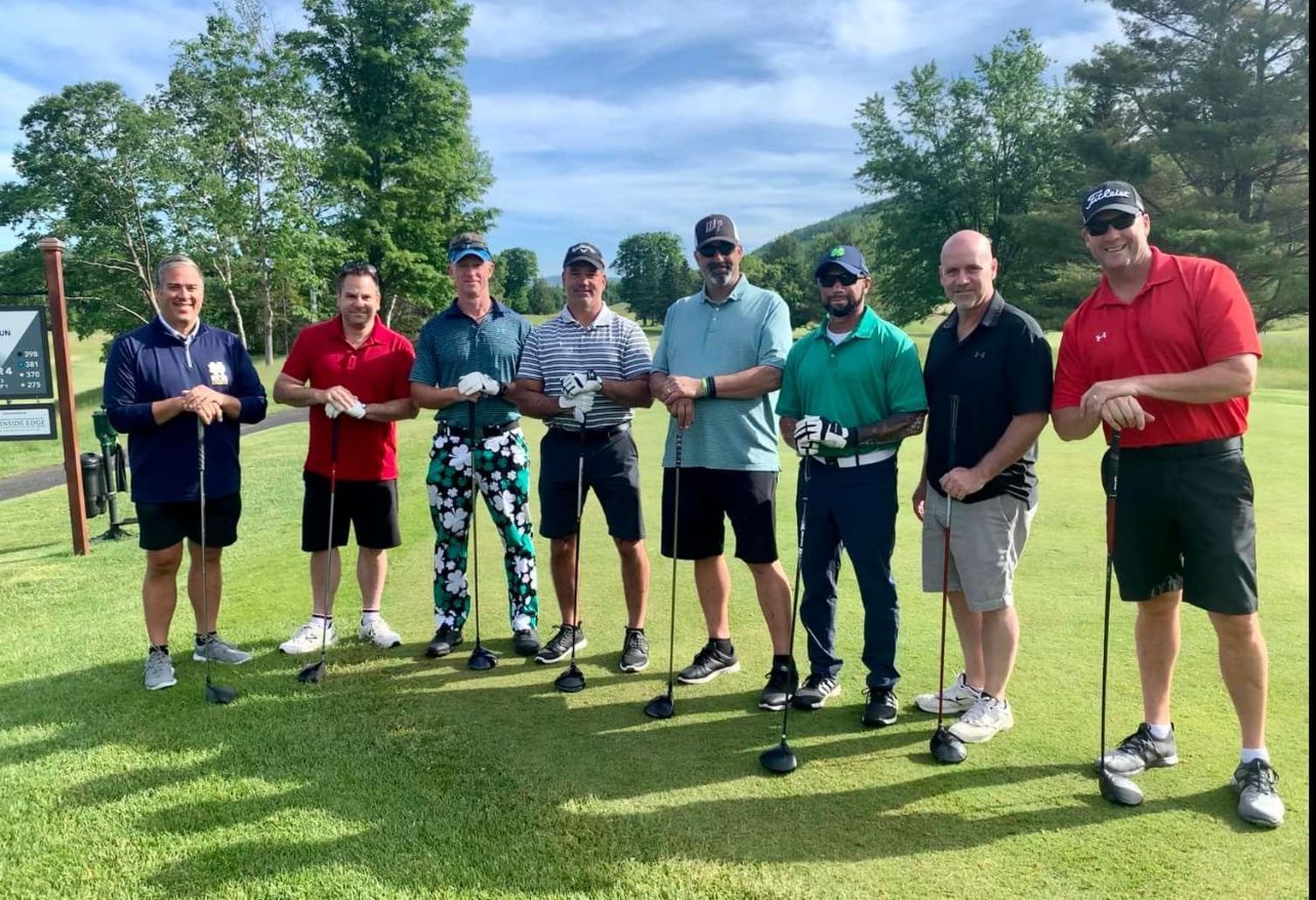 Group of men standing on a golf course, holding clubs. They wear casual clothes.