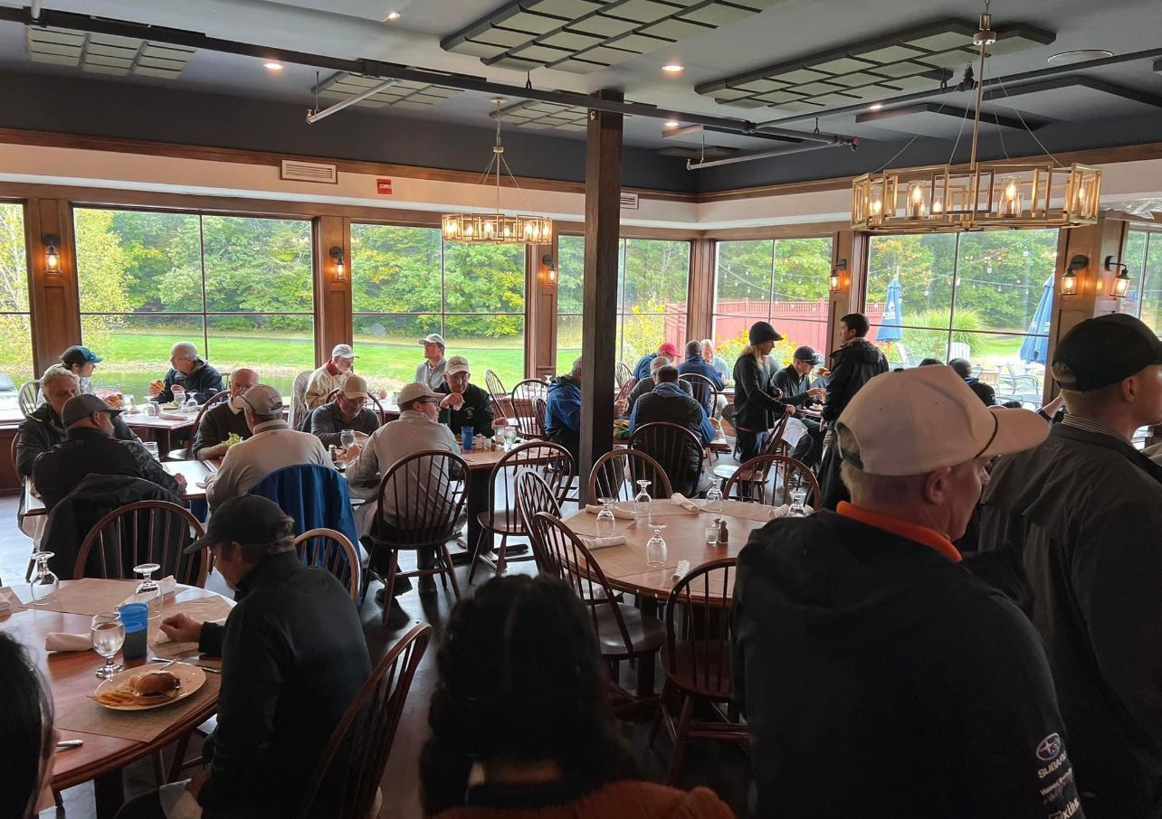 Restaurant interior with patrons seated at tables near large windows overlooking a green space.