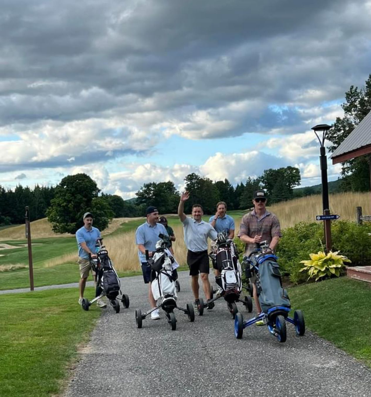 Five men on a golf course, pushing golf bags on carts. One waves, cloudy sky.