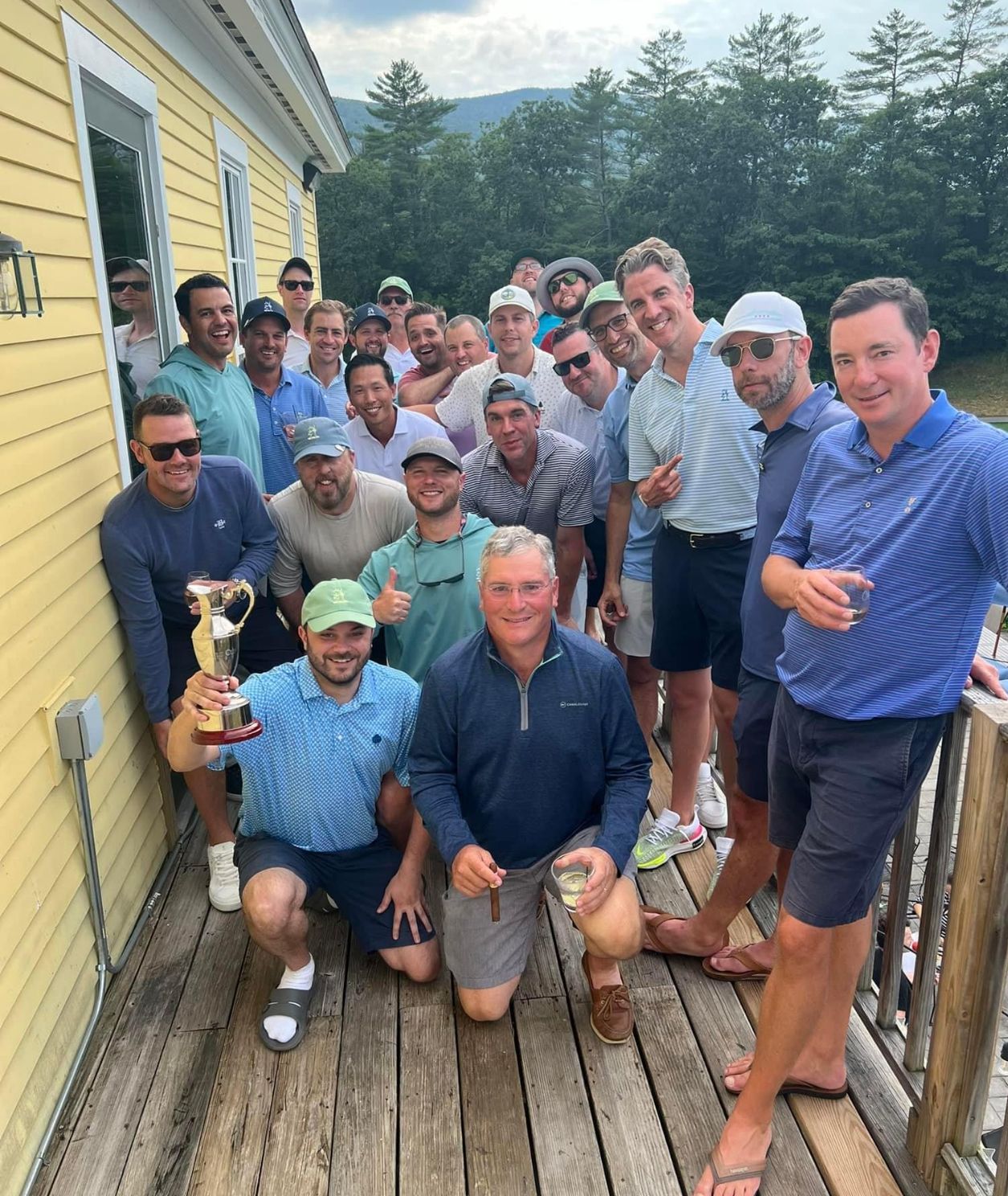 Group of men pose on a deck with a trophy; outdoors, mountain backdrop. Smiling, celebrating.