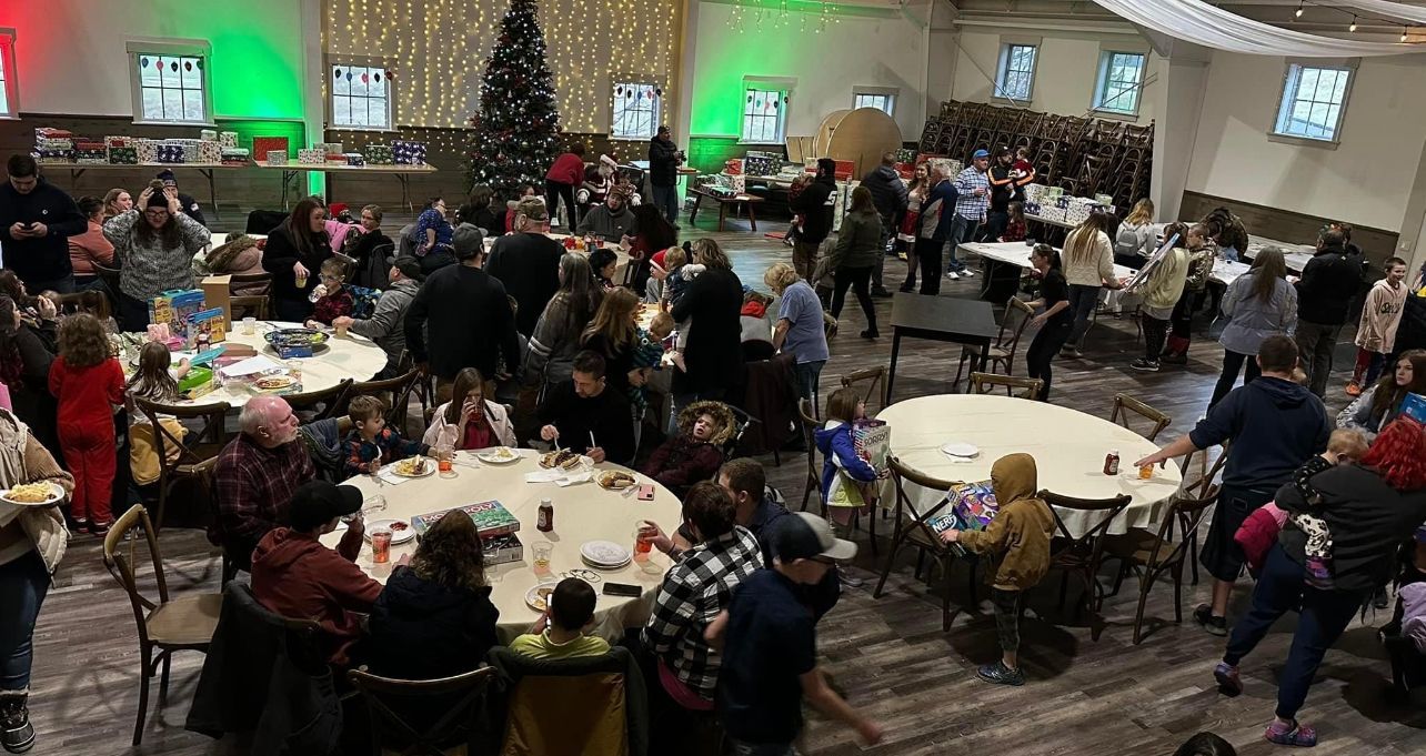 Large room with holiday party, people gathered around tables, Christmas tree, green and red lights.