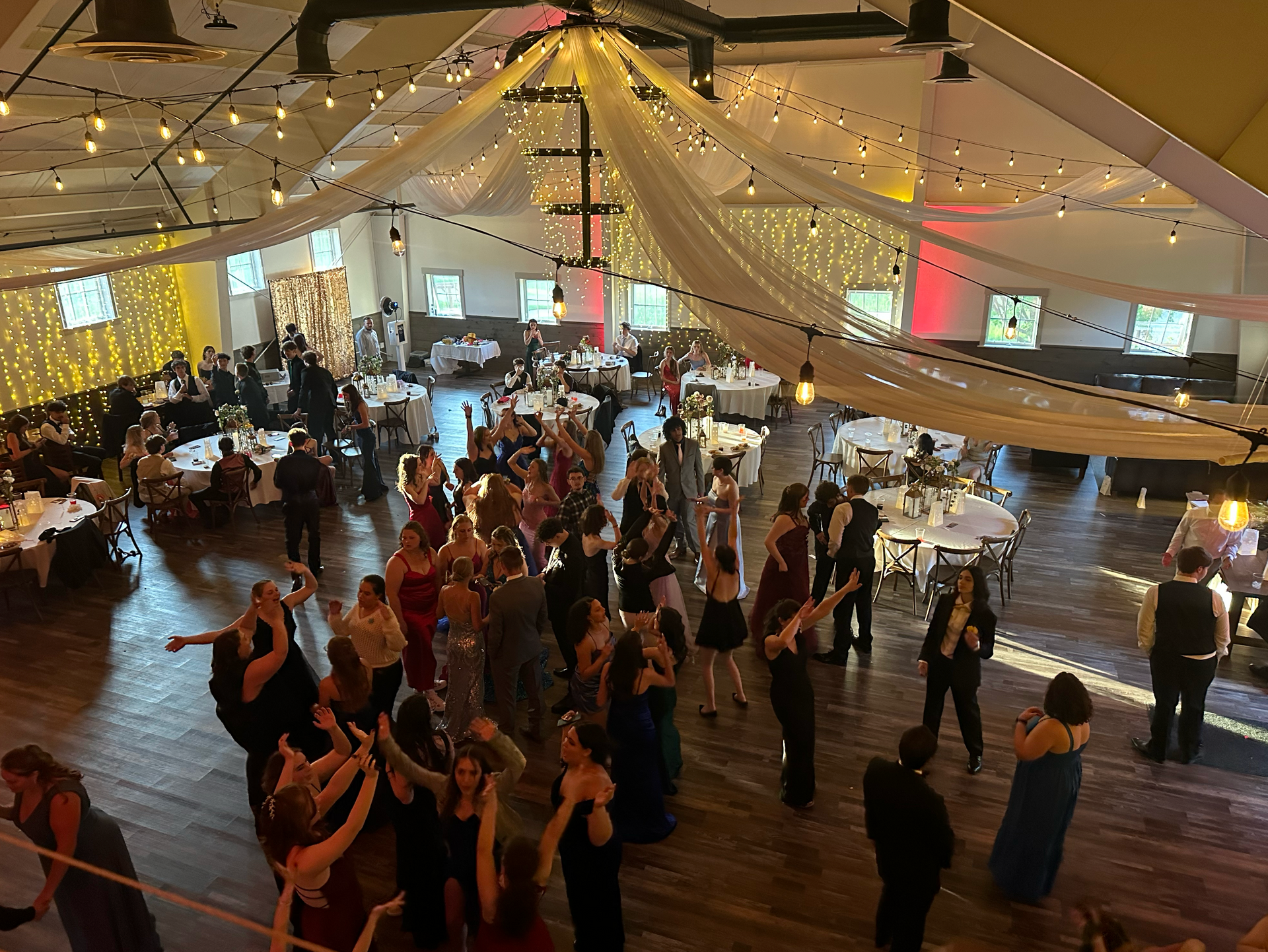 People dance at a formal event in a large, decorated hall with string lights and draped fabric.