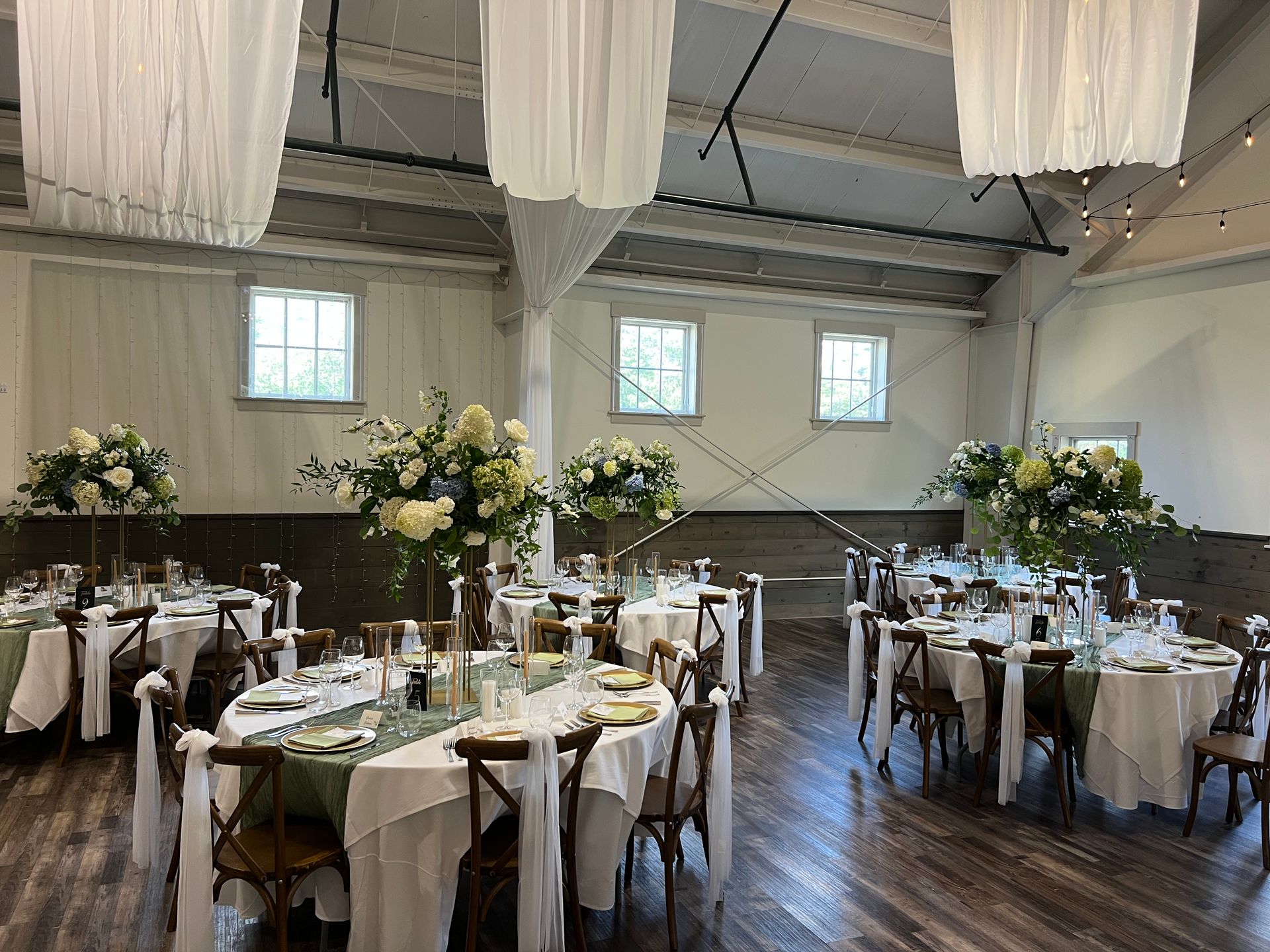 Wedding reception: round tables with white linens, floral centerpieces, and cross-back chairs in a bright room.