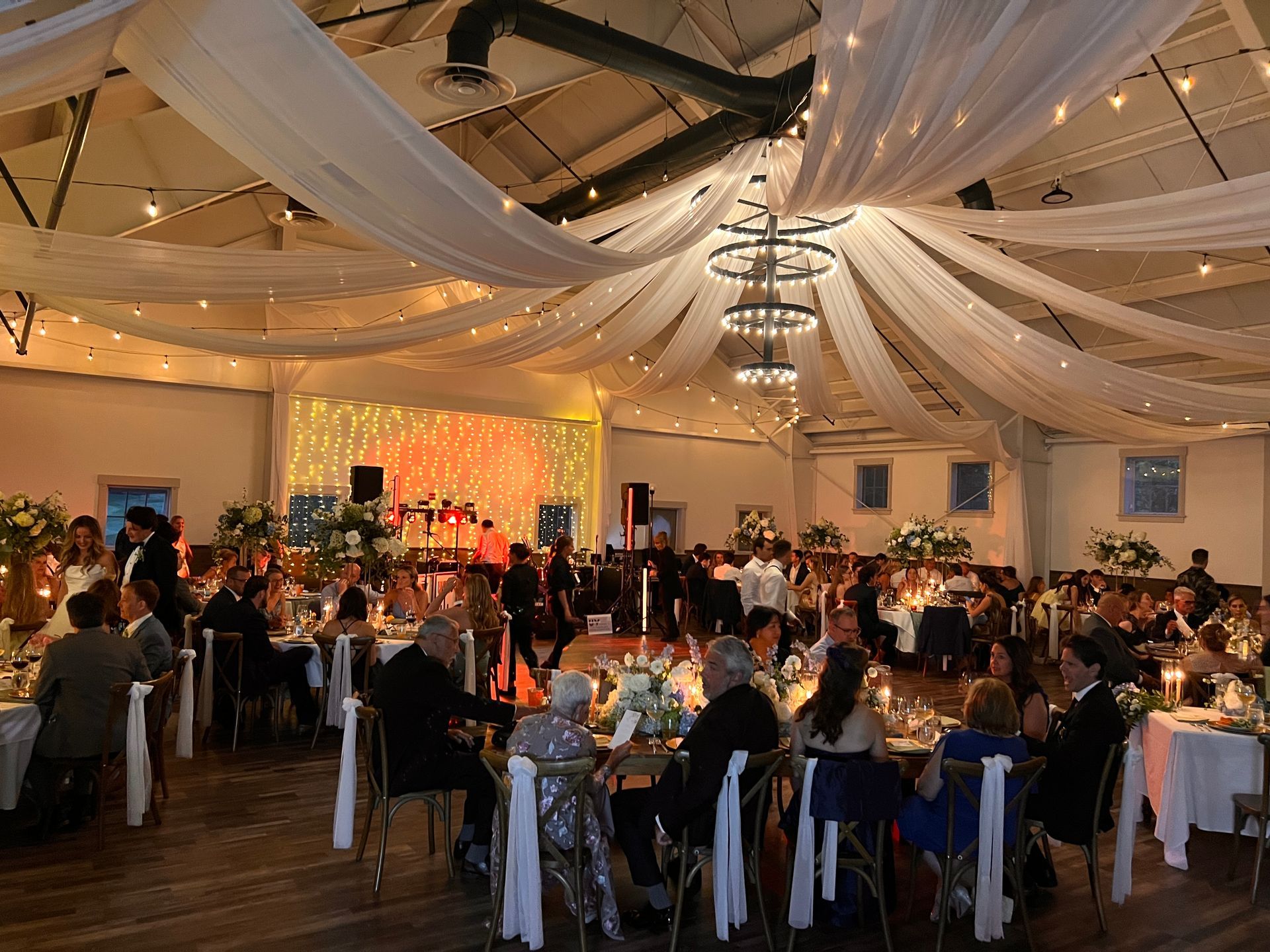 Elegant banquet hall, draped ceiling with soft lighting, guests seated at tables, lively atmosphere.