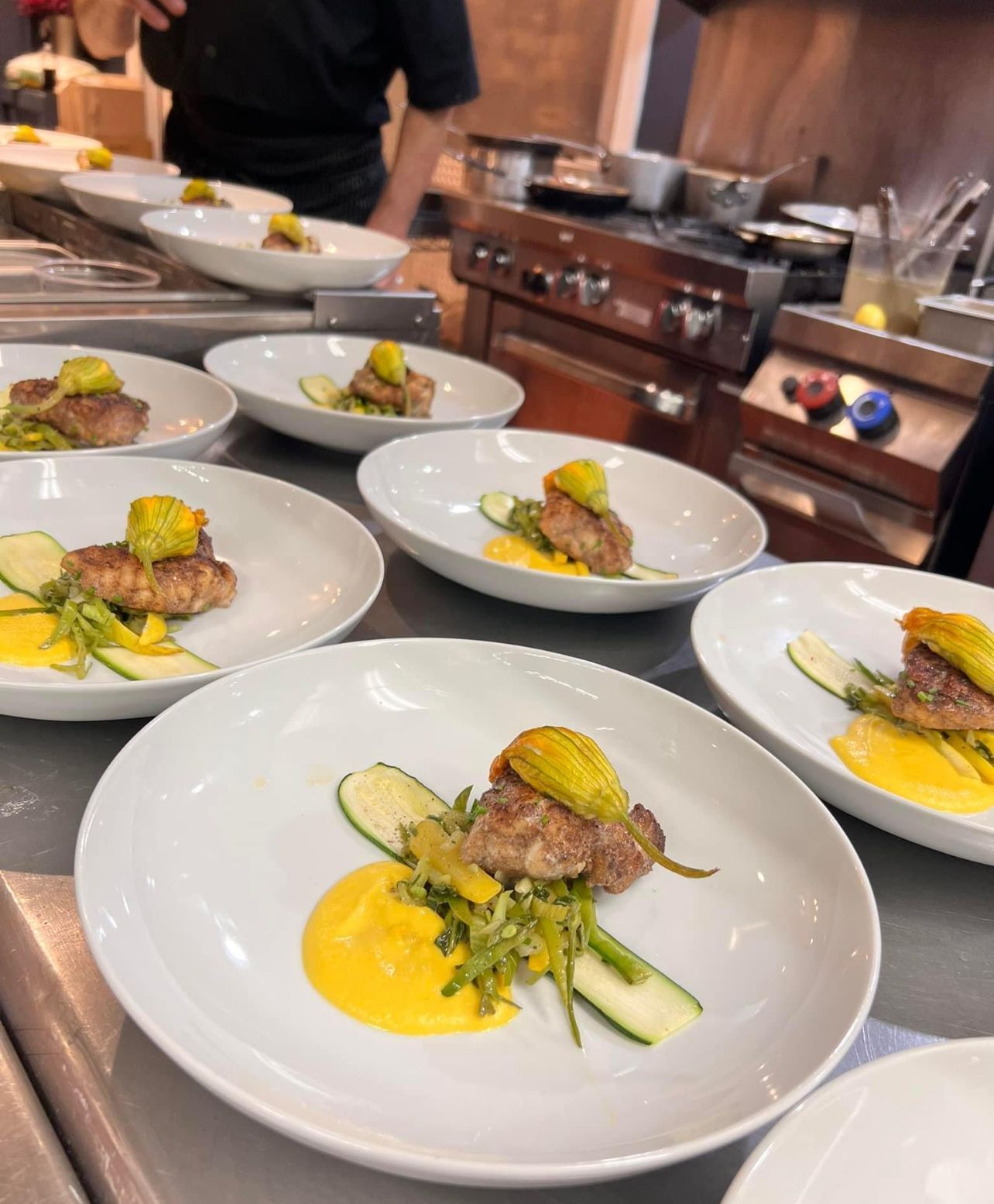 Plated dishes with seared meat, yellow sauce, zucchini, and squash blossoms, on a kitchen counter.