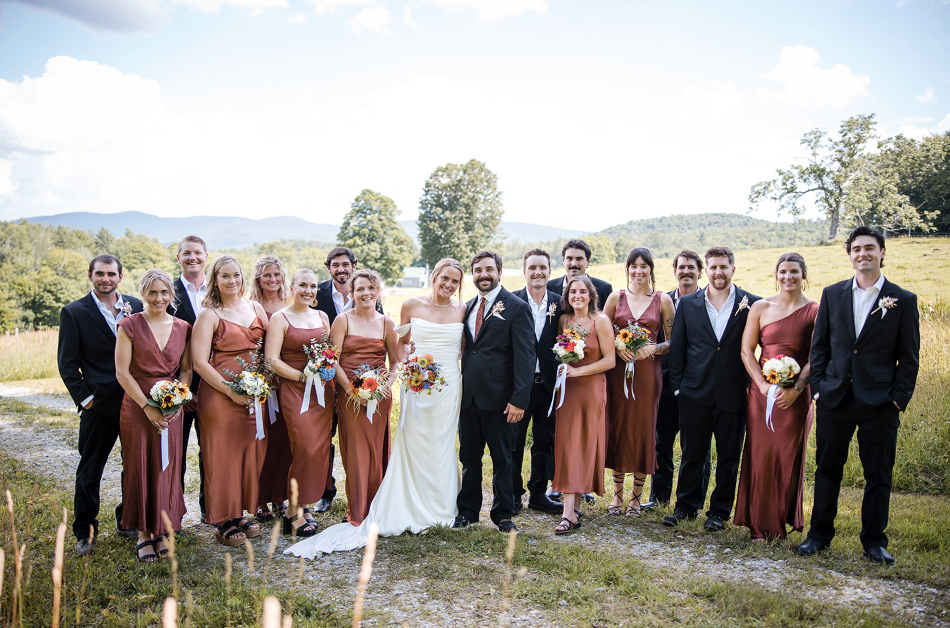 Wedding party poses outdoors. Bride and groom center. Bridesmaids in rust, groomsmen in black suits. Green hills in background.