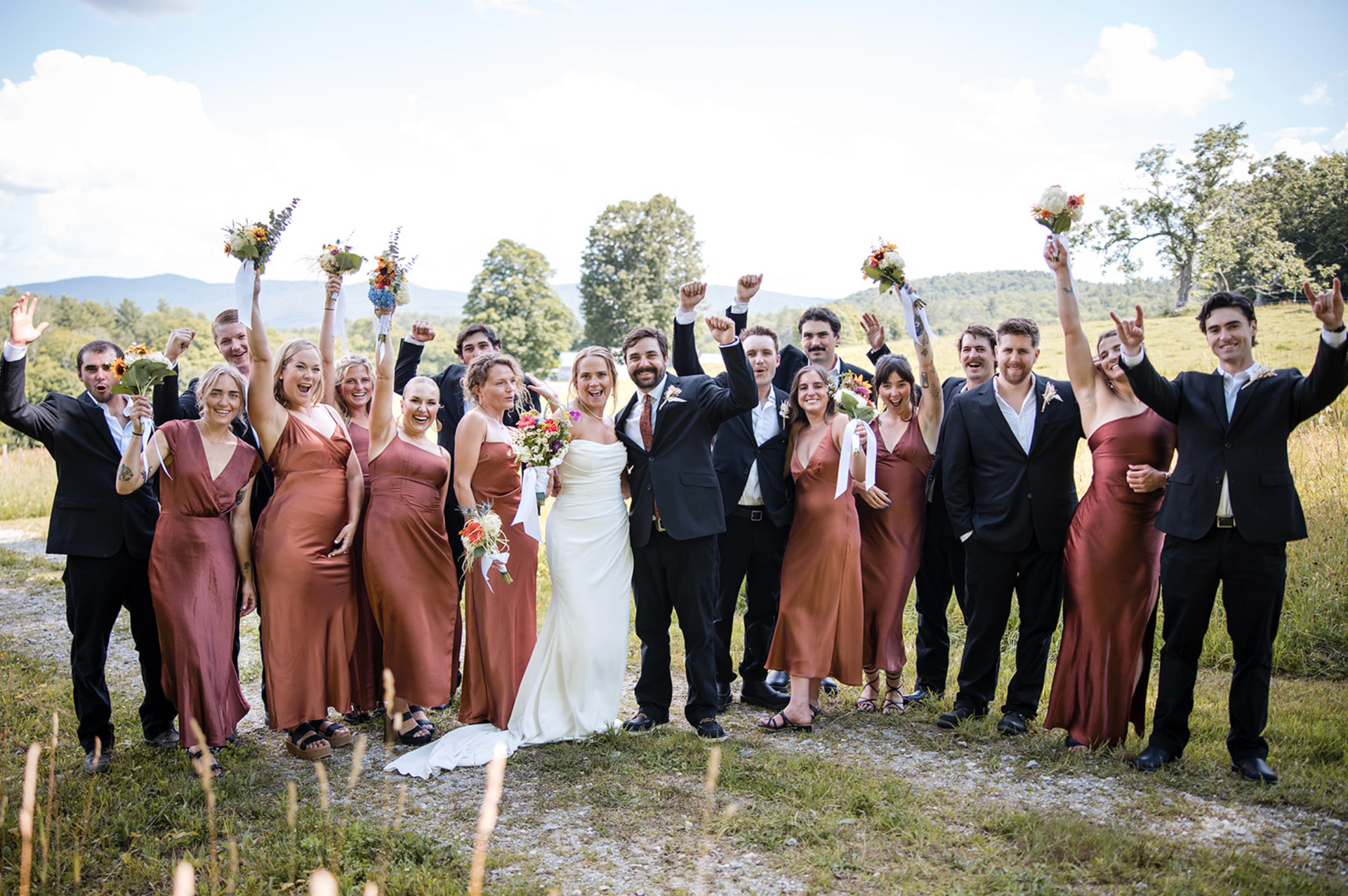 Wedding party celebrates outdoors, arms raised. Bride & groom in center, surrounded by bridesmaids in rust-colored dresses and groomsmen in black suits.