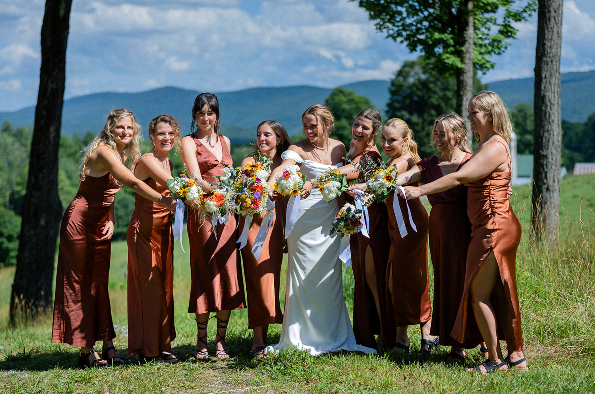 Bridesmaids in rust-colored dresses and bride in white dress hold bouquets. Mountain backdrop on a sunny day.