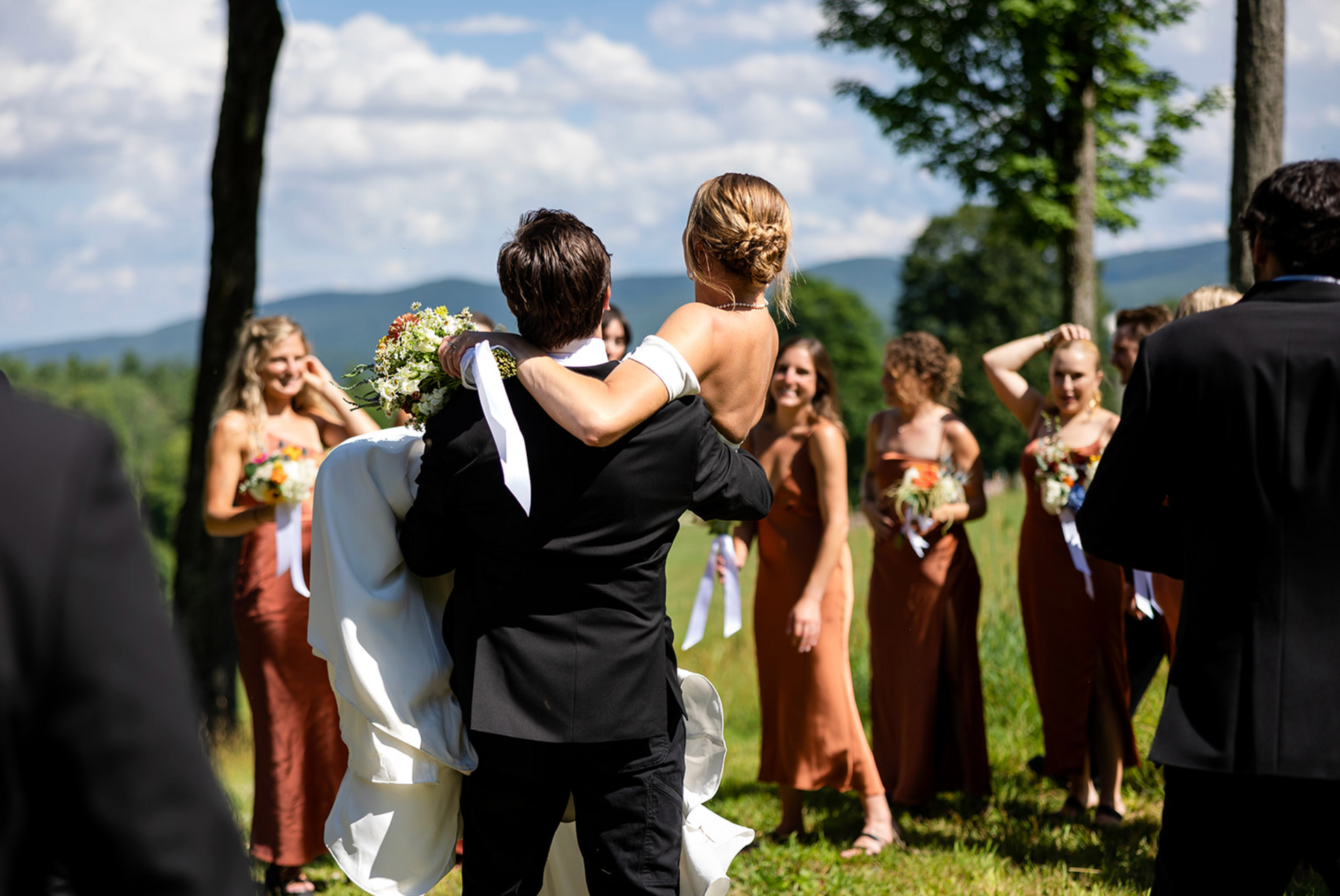 Groom lifts bride during wedding ceremony. Bridesmaids in orange dresses watch. Mountain view background.