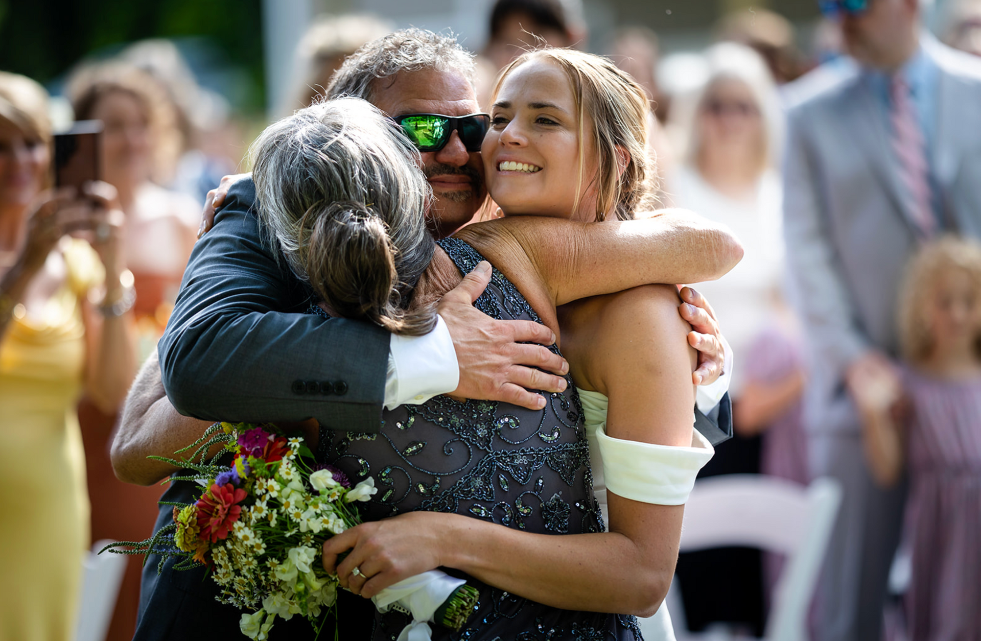 Father hugs bride at outdoor wedding, both smiling; guests watch.