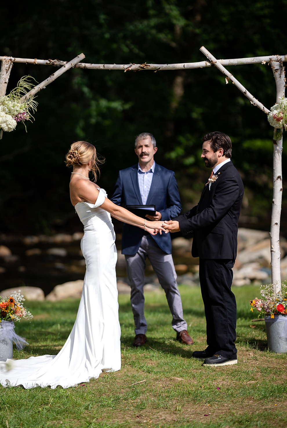 Bride and groom hold hands during outdoor wedding ceremony; officiant stands nearby under rustic arch.