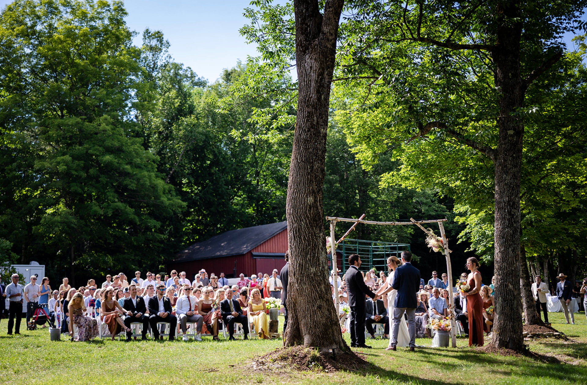 Wedding ceremony outdoors with guests seated, officiant and couple under an arbor. Green trees and a red barn.