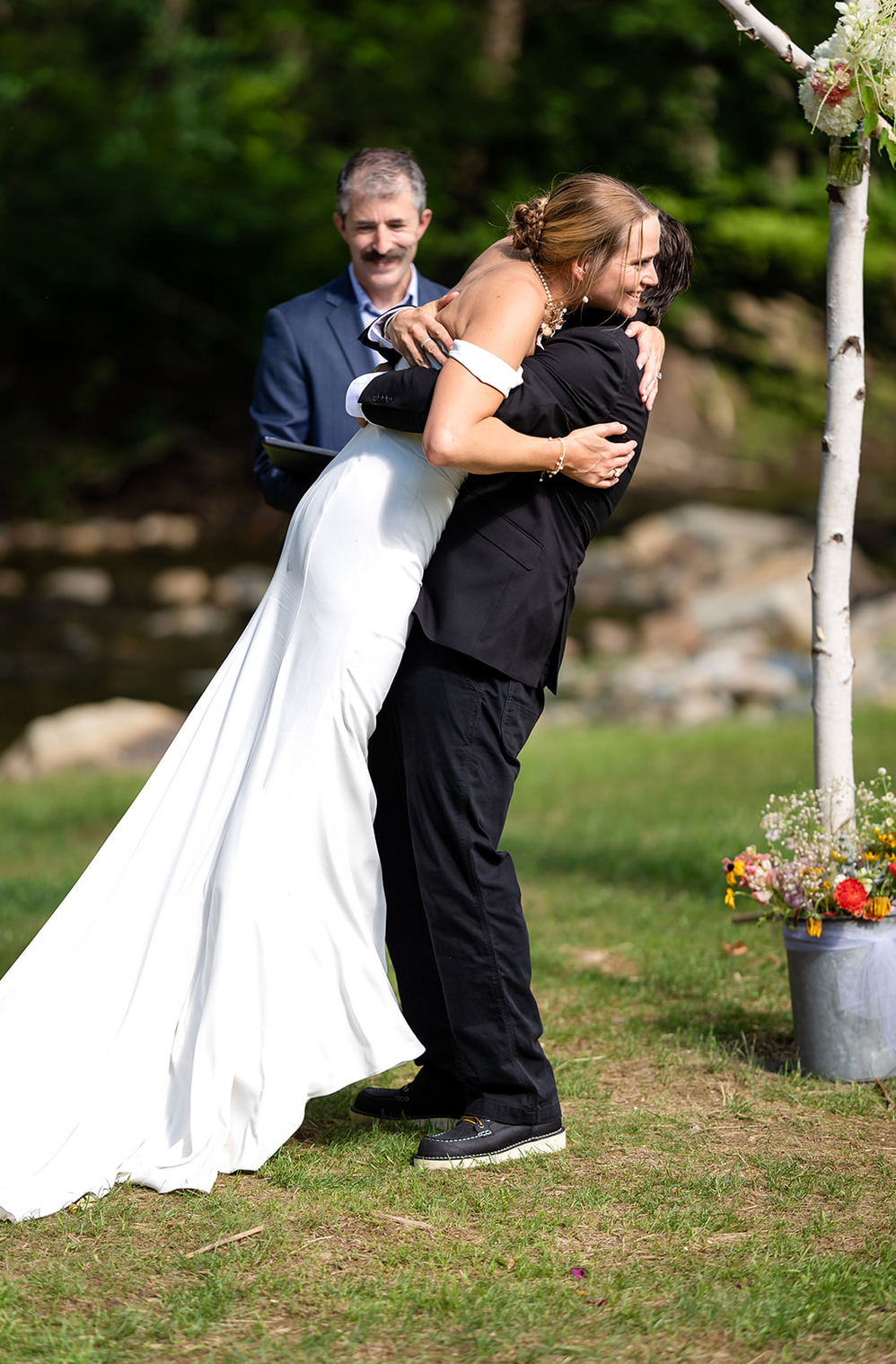 Groom hugs bride after wedding ceremony, white dress, black suit, officiant in background. Outdoor setting, green grass.