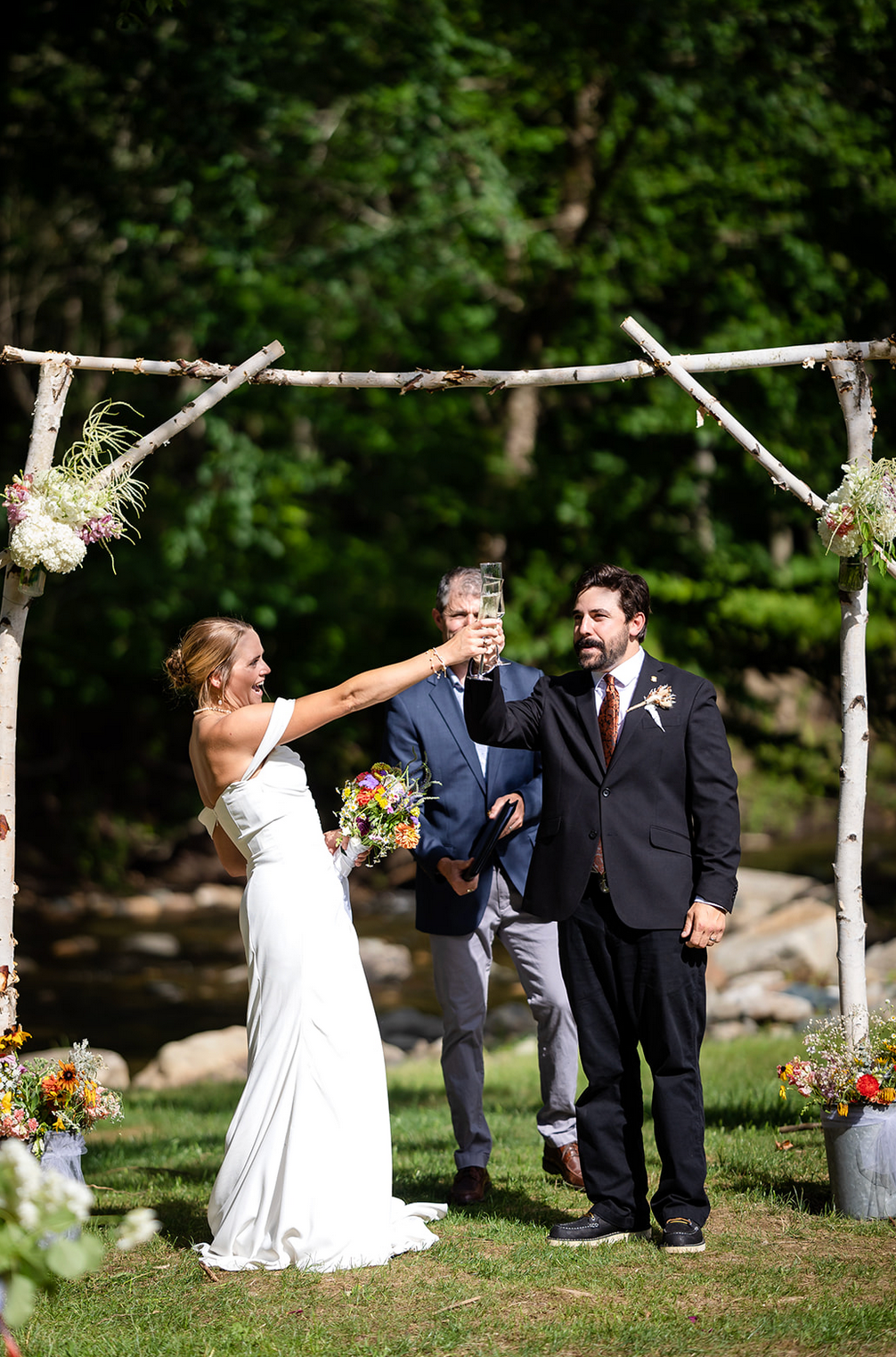 Bride and groom toast at outdoor wedding under birch arch. Bride laughs, groom smiles; officiant stands behind.