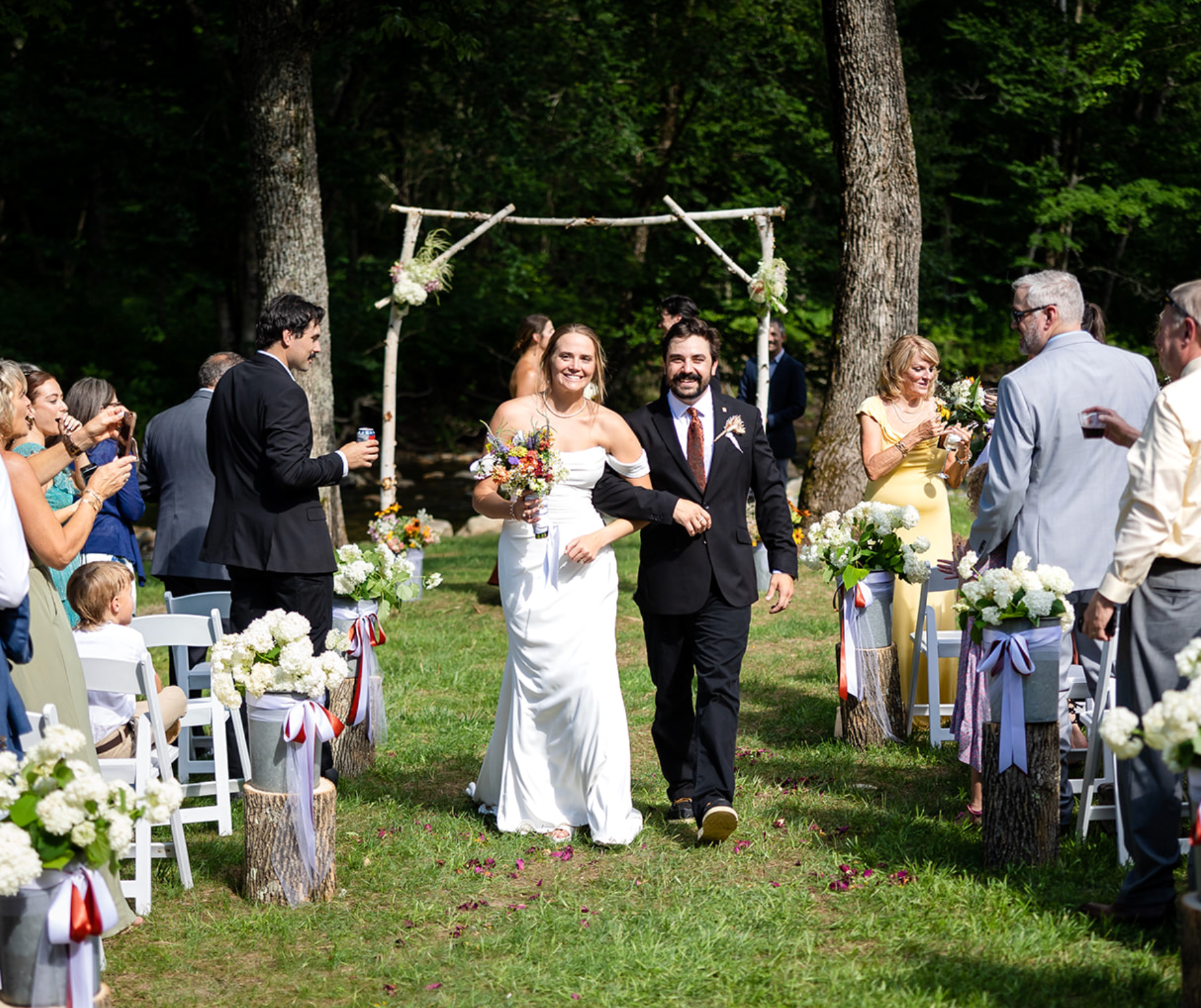 Newlyweds walk down the aisle after their wedding ceremony in a forest, cheered on by guests.