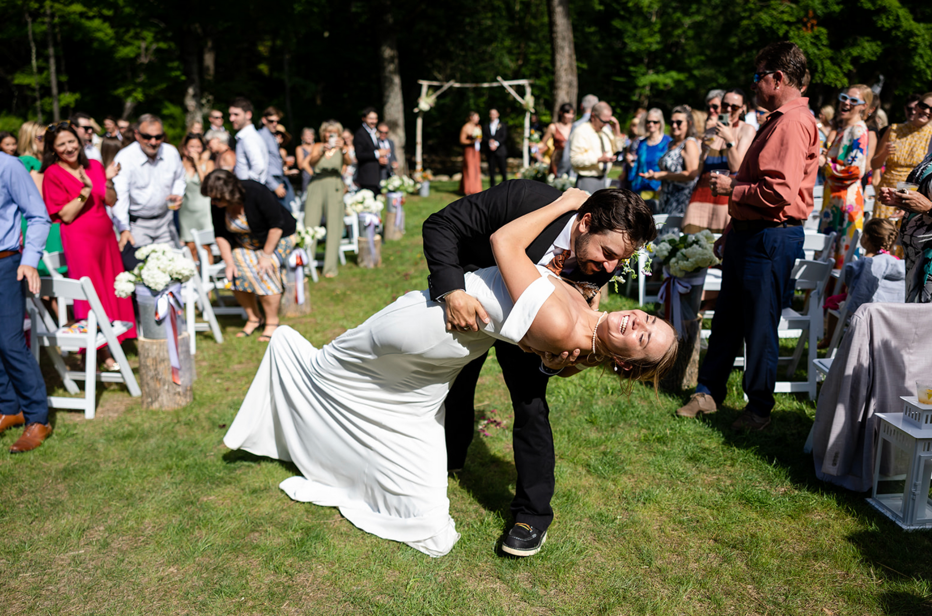 Newlyweds dip during outdoor wedding ceremony, surrounded by cheering guests in a grassy setting.
