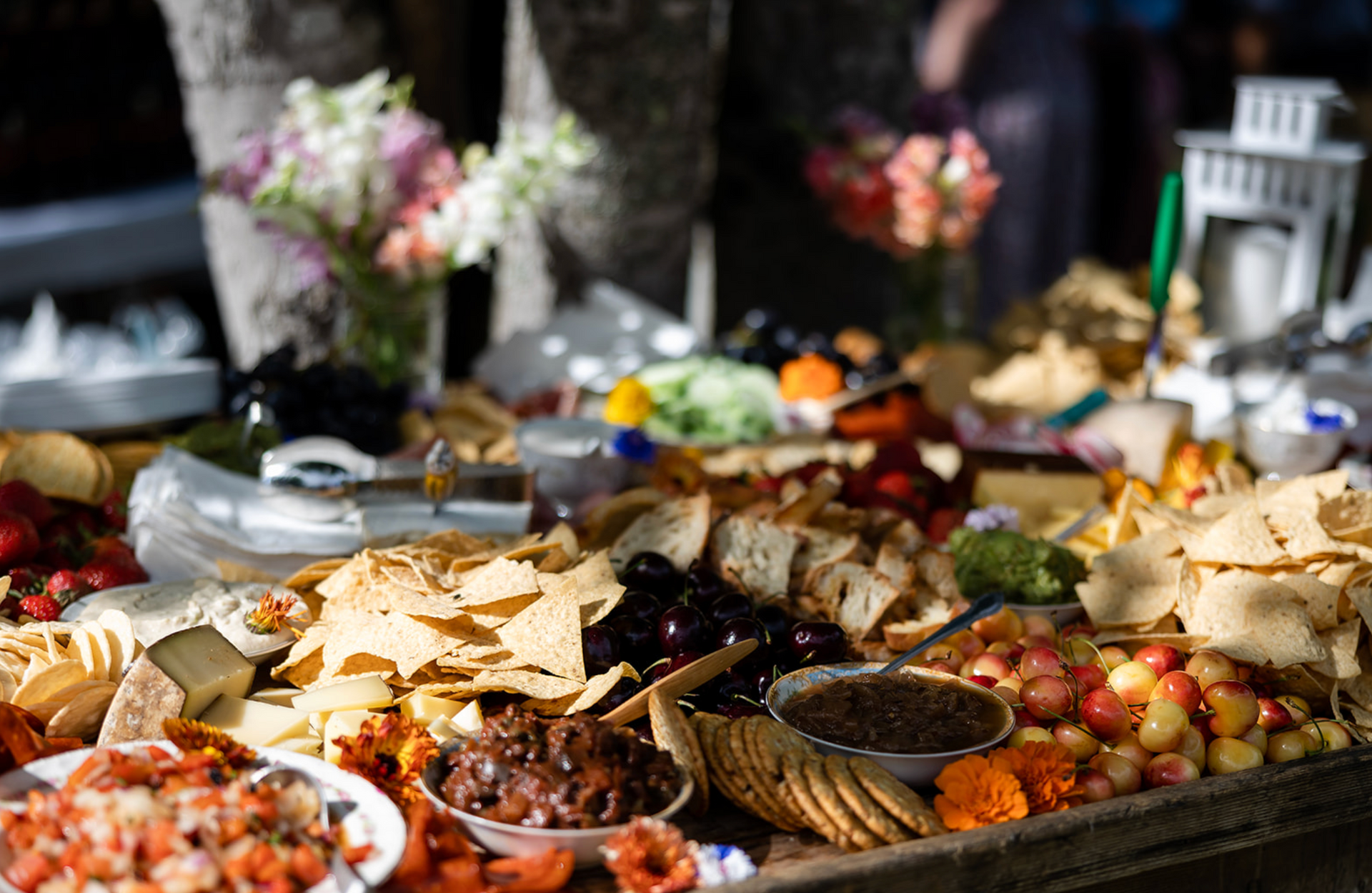 A buffet table laden with food, including chips, dips, cheeses, and crackers, with flower arrangements in the background.