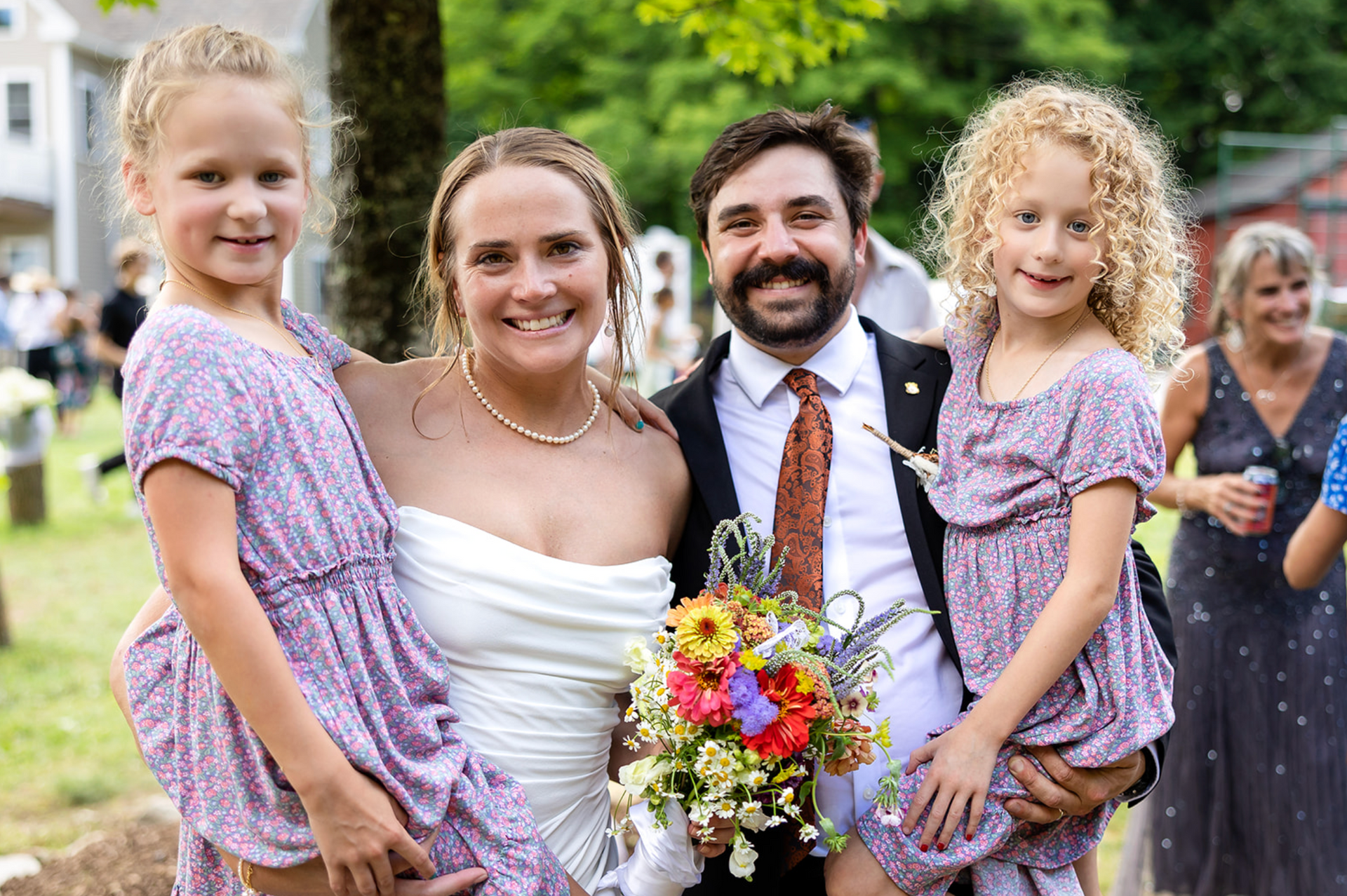 Newly married couple with two young girls wearing matching dresses, smiling outdoors.
