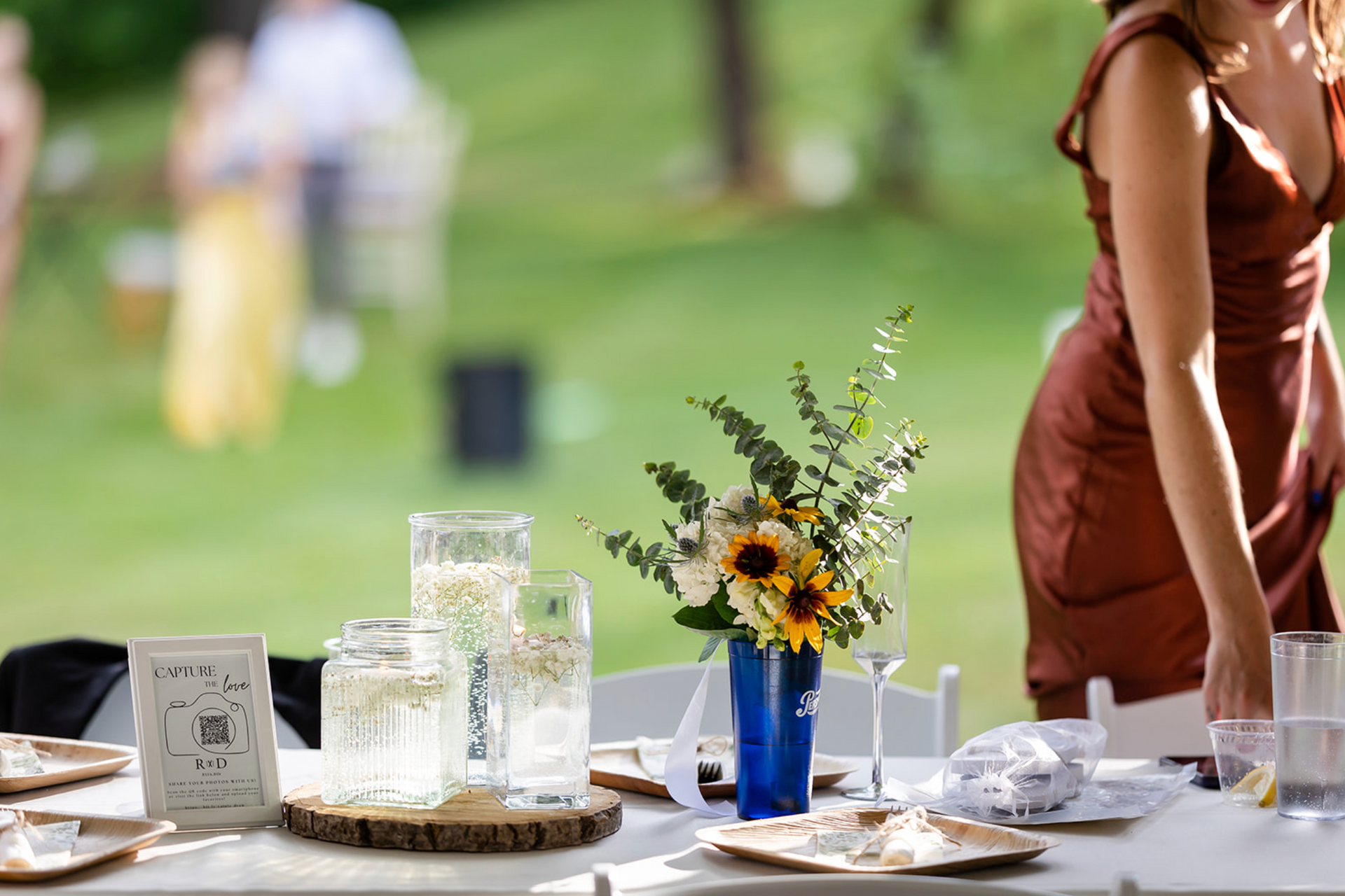 A woman adjusts a table setting outdoors, featuring sunflowers, water glasses, and plates.