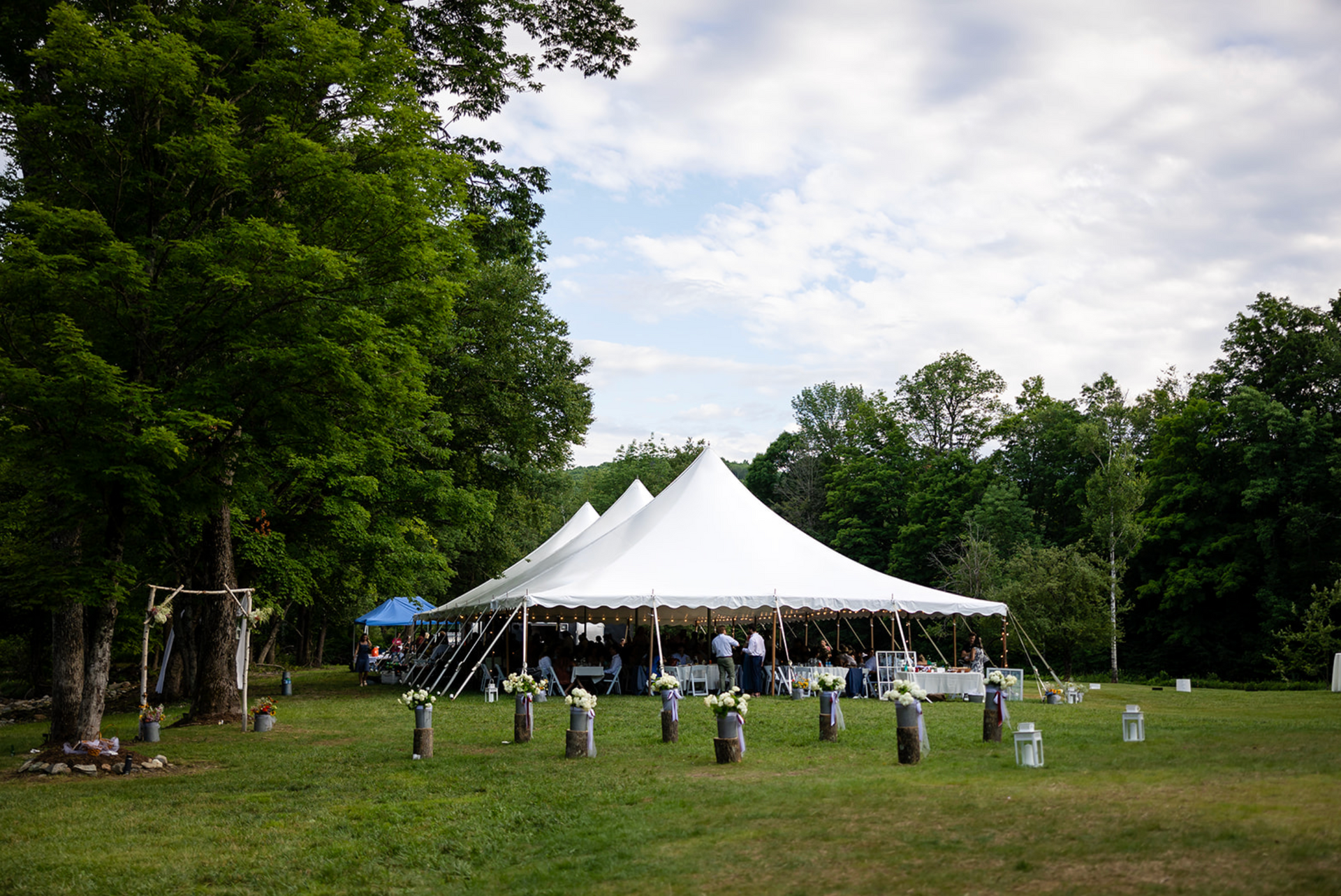 White tent set up on green grass for an event, surrounded by trees and flower decorations.