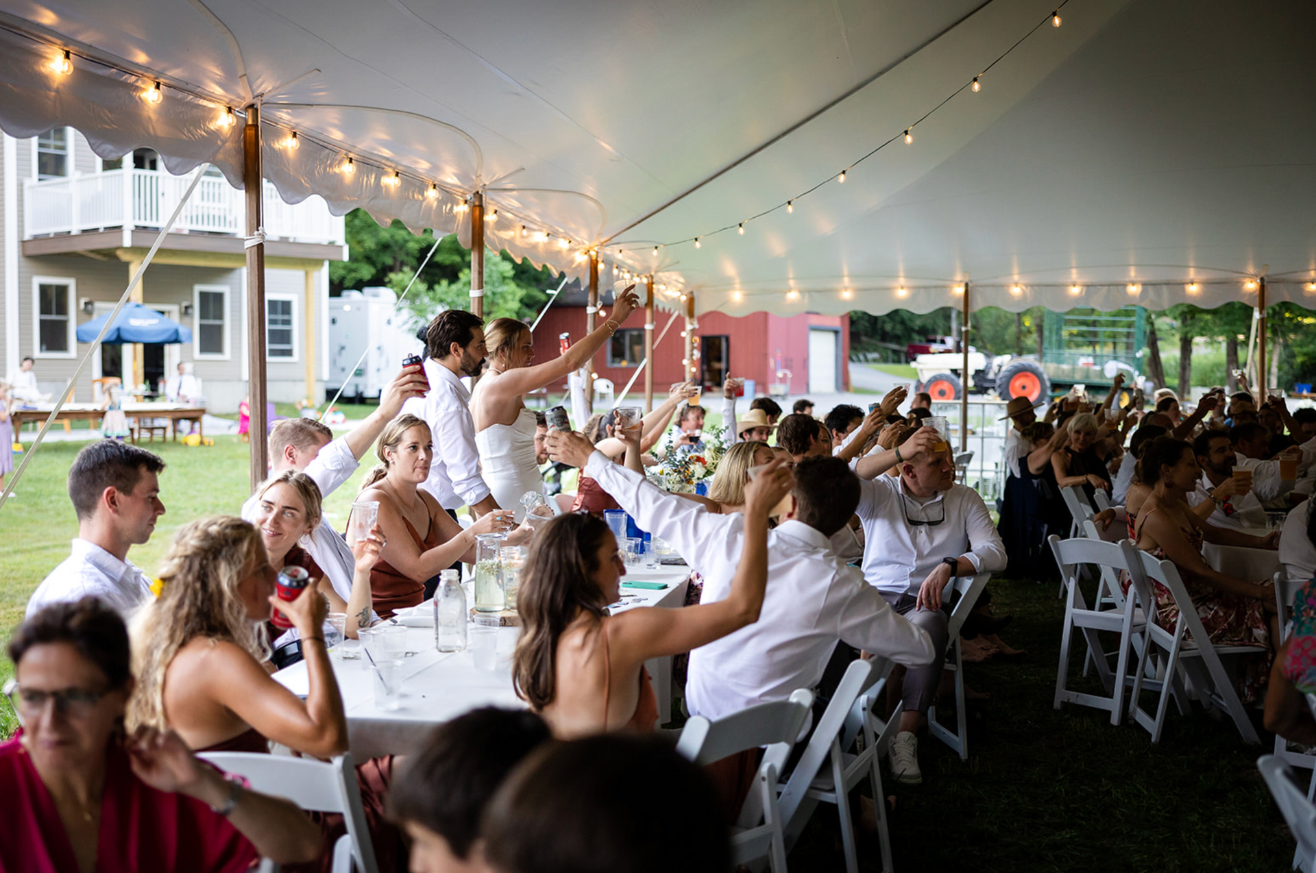 Wedding reception under a white tent; guests raise glasses and cheer at tables; string lights overhead.