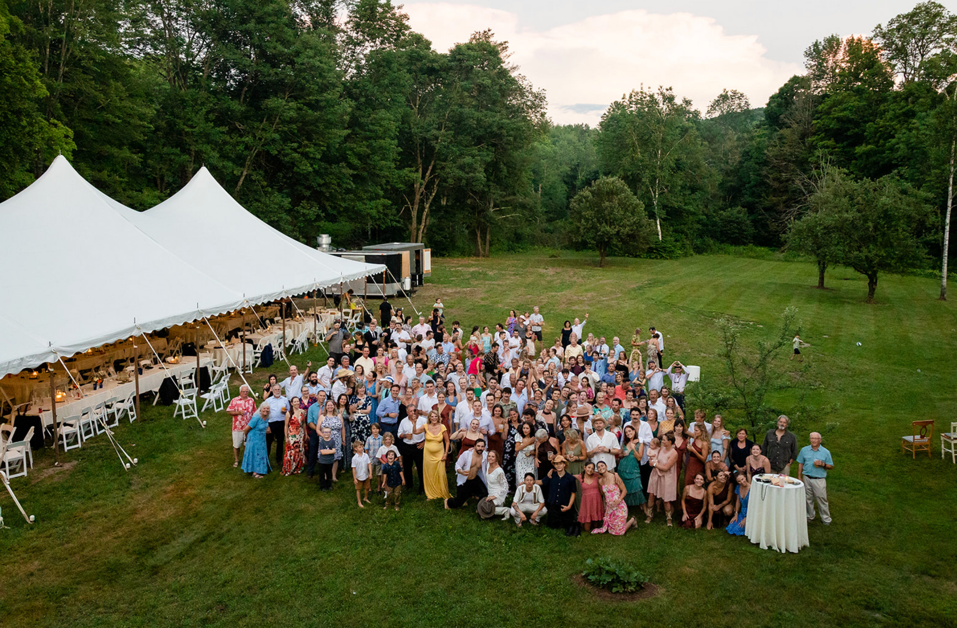 Large group of people gathered outdoors for an event near a white tent, surrounded by trees.