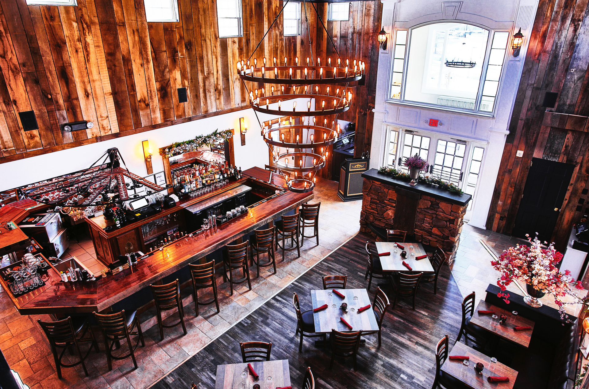 Interior of a rustic restaurant with a bar, tables, and a large chandelier. Wooden walls and floors.