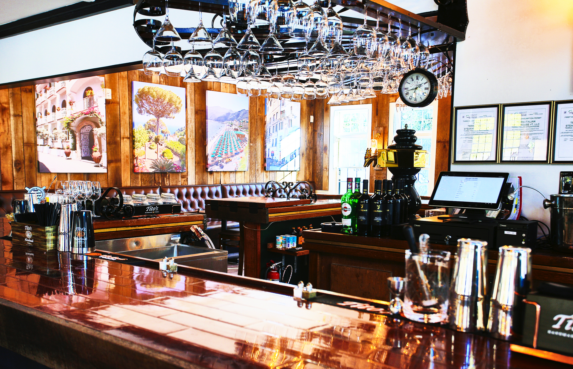 Bar interior with copper counter, wooden paneling, paintings, hanging glasses, and a clock.