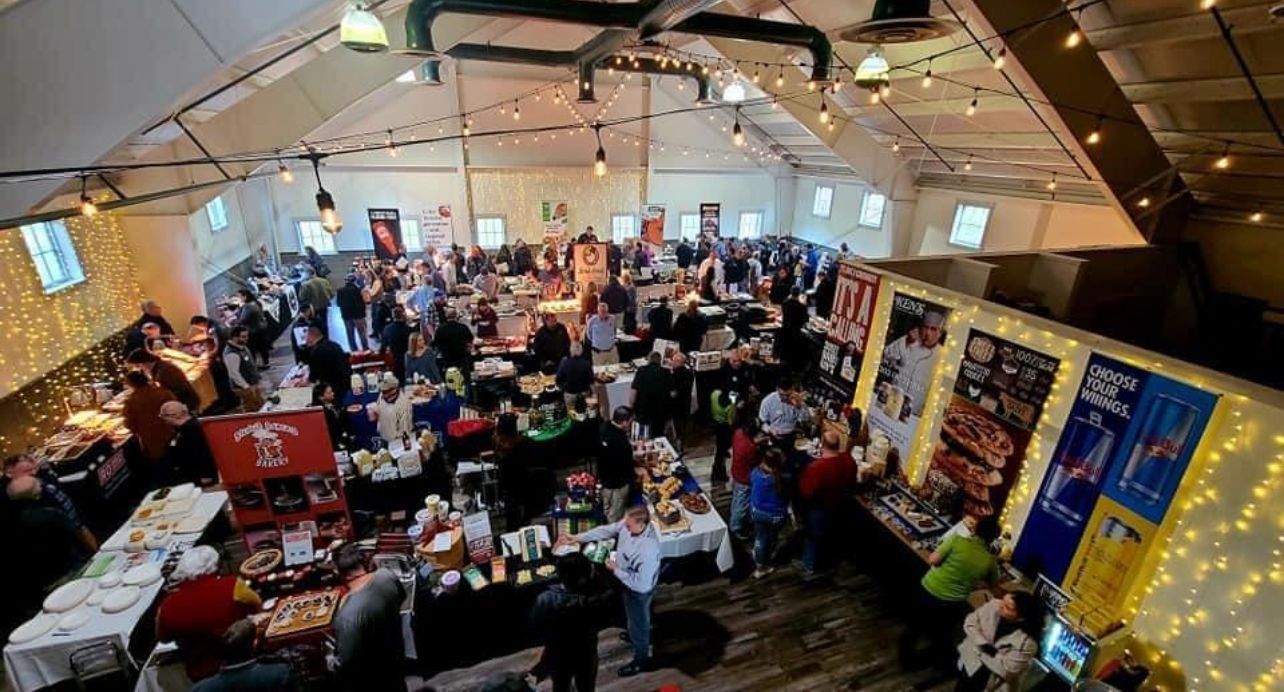 Large event space packed with people browsing vendor tables; warm lighting, festive atmosphere.