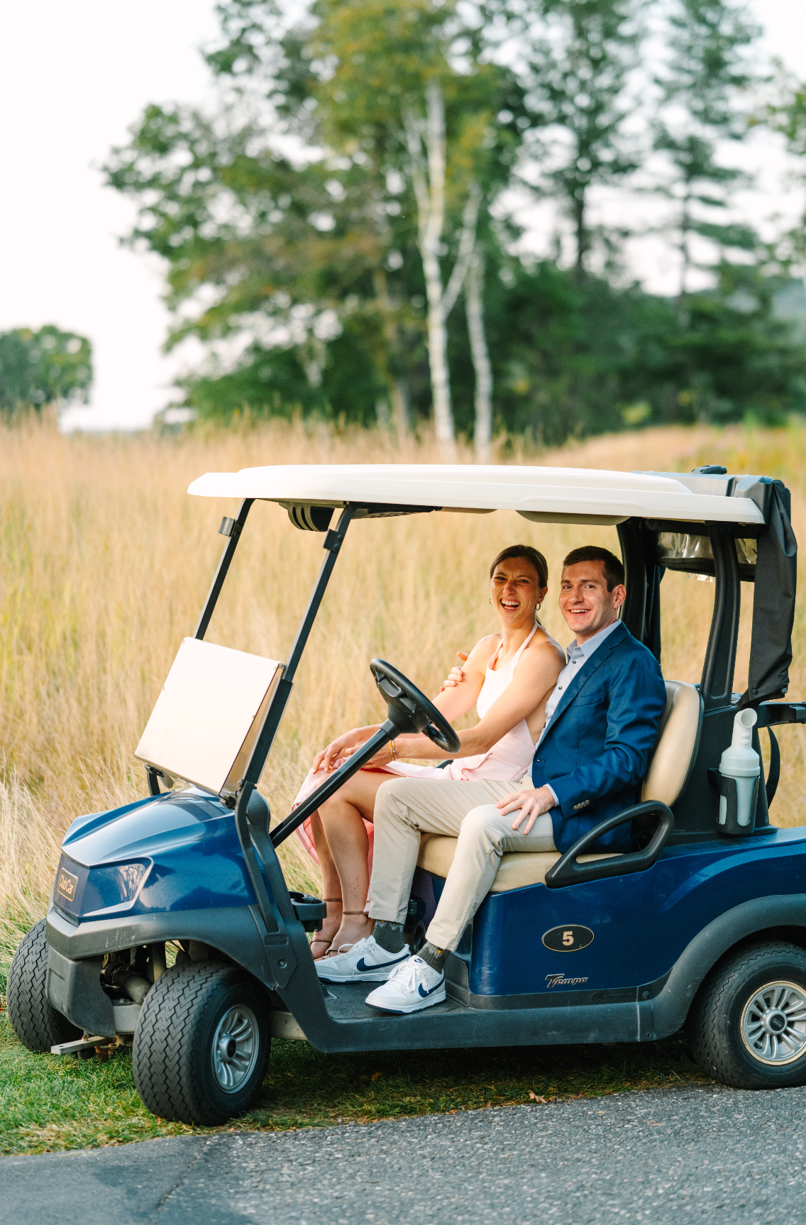 Couple in a blue golf cart, smiling, set against a field of tall yellow grass.