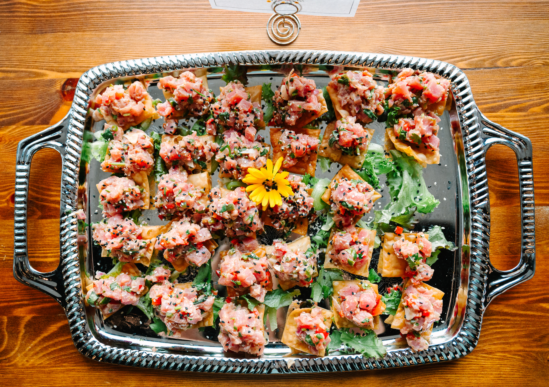 Silver tray of tuna tartare appetizers on wooden table, garnished with a yellow flower.