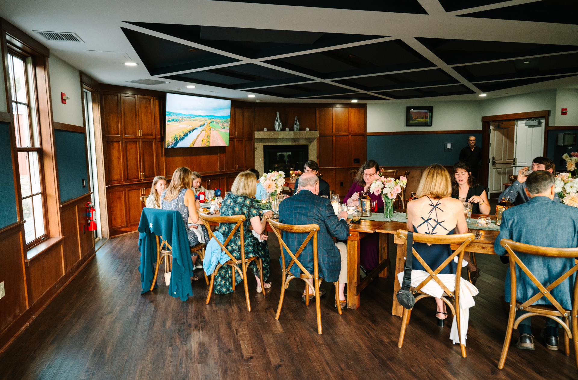 People seated at a long table in a wood-paneled room. Guests are dining, and the room has a fireplace and a TV.