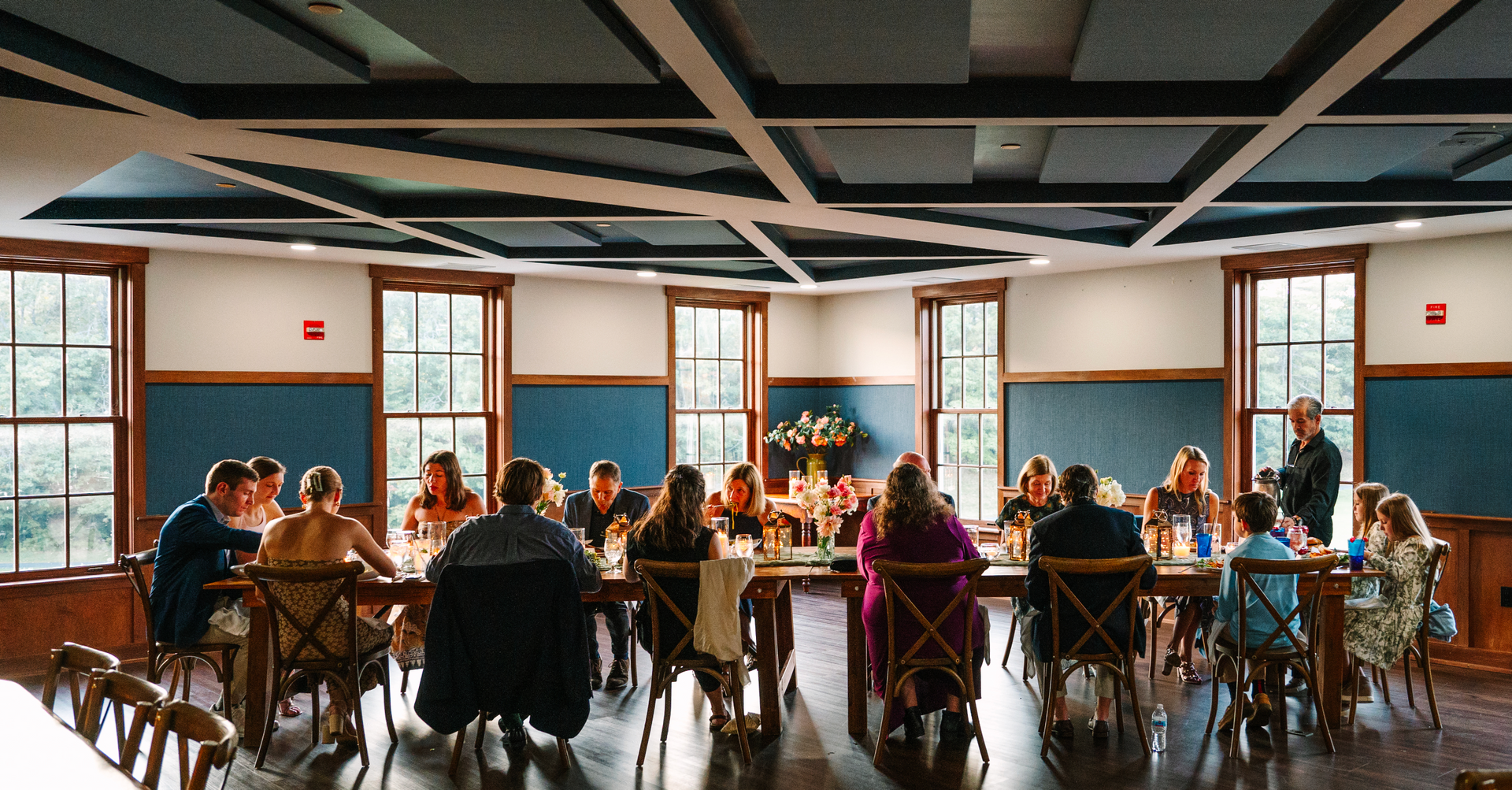 Large group seated at a long table. People are dining in a well-lit room with windows and a geometric ceiling.