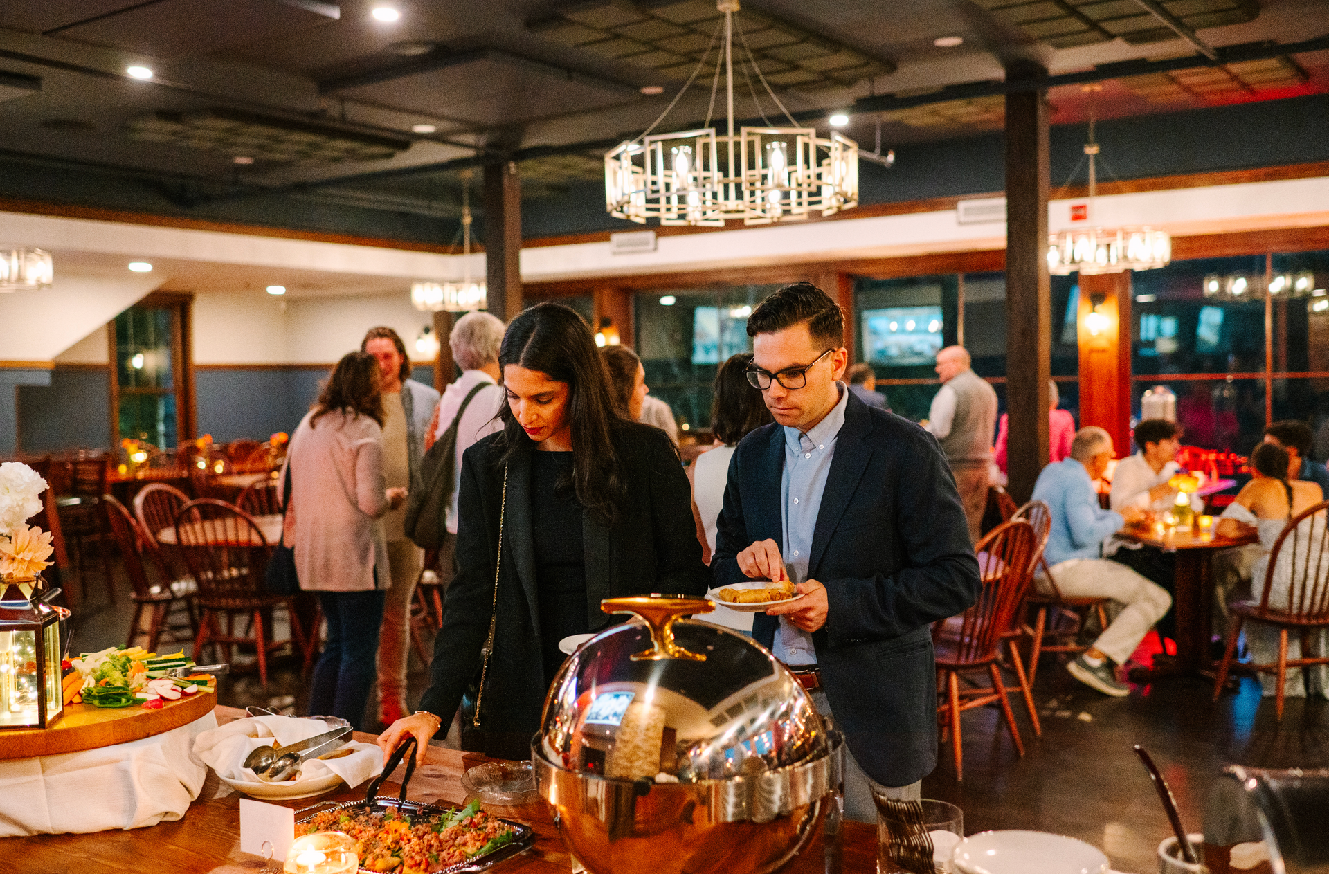 People at a buffet in a warmly lit restaurant, selecting food from serving dishes.