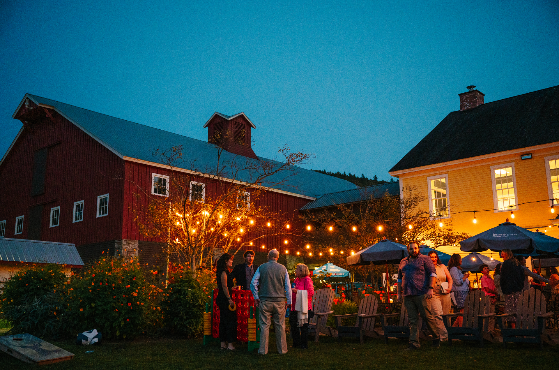 Evening outdoor event with people gathered near a red barn and yellow building, string lights overhead.