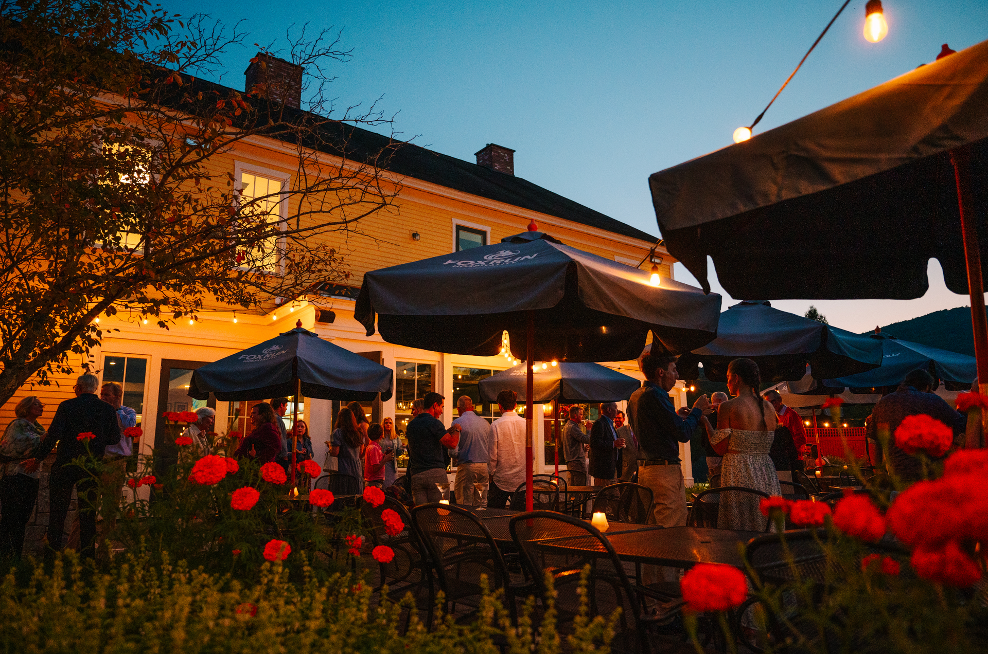 People gather outside a yellow building at dusk, under umbrellas with string lights. Red flowers in foreground.