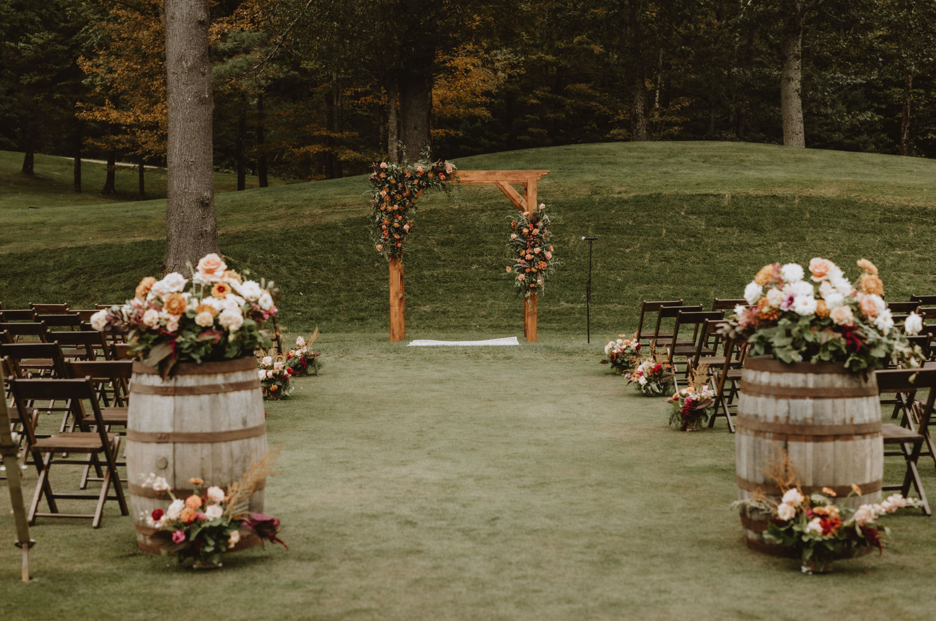 A wedding ceremony is taking place in a field with wooden barrels filled with flowers.