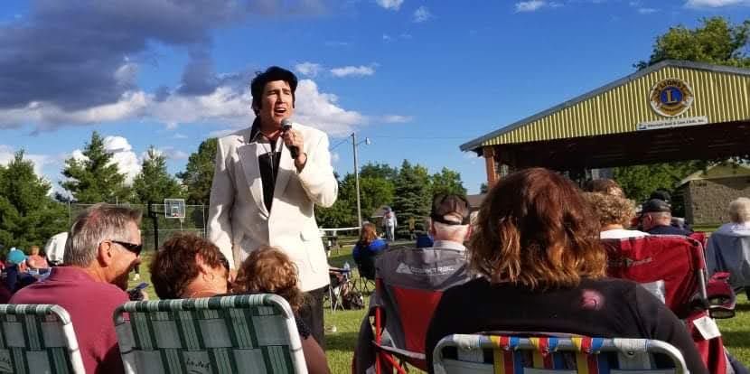 Man speaking into a microphone to an outdoor seated crowd near a pavilion on a sunny day