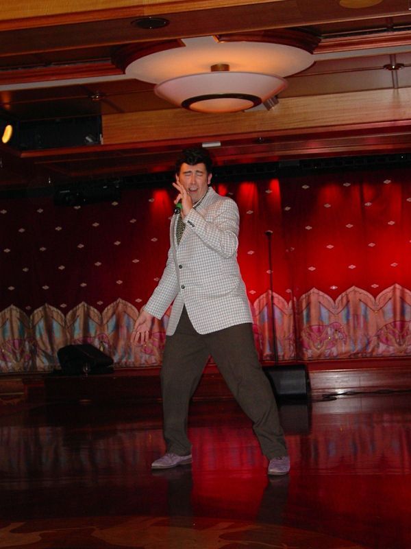 Man posing on a red-lit stage with a microphone, wearing a light jacket and dark pants.