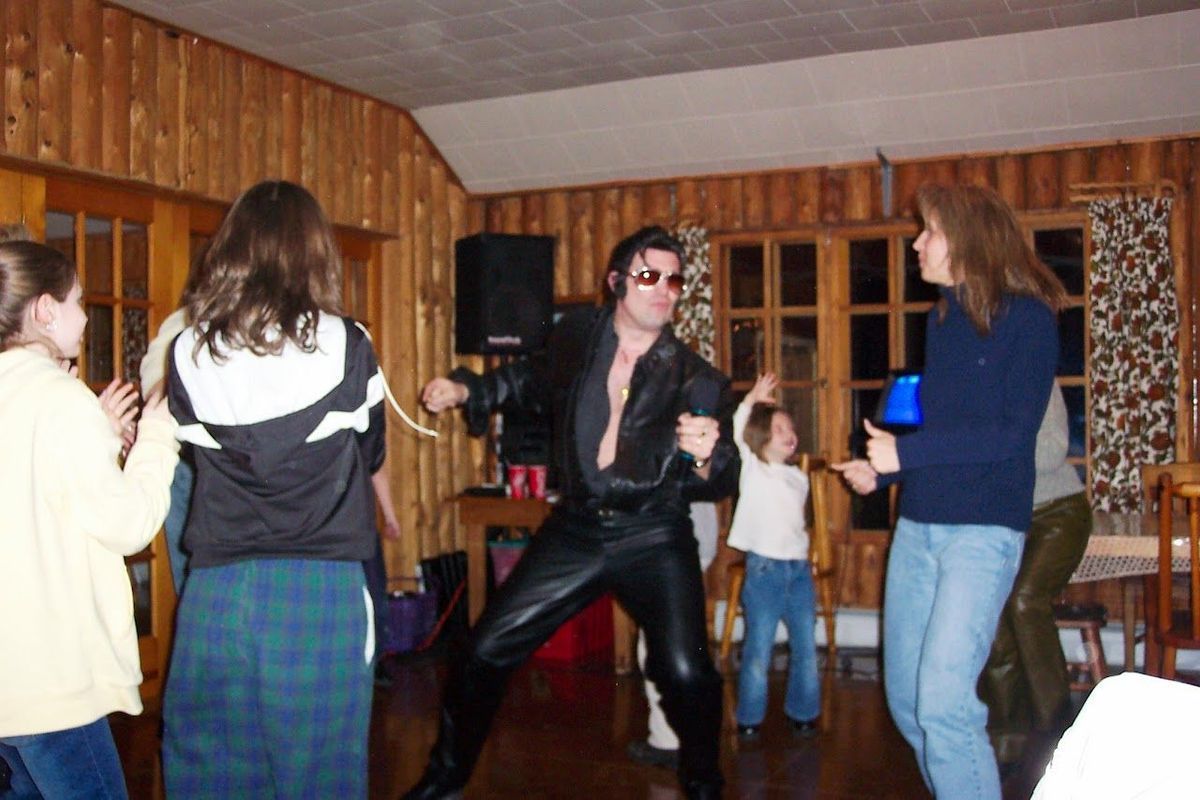 Person in black leather dancing in a wood-paneled room while others watch at a party