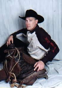 Man in black cowboy hat and embroidered jacket sitting with a saddle against a light backdrop